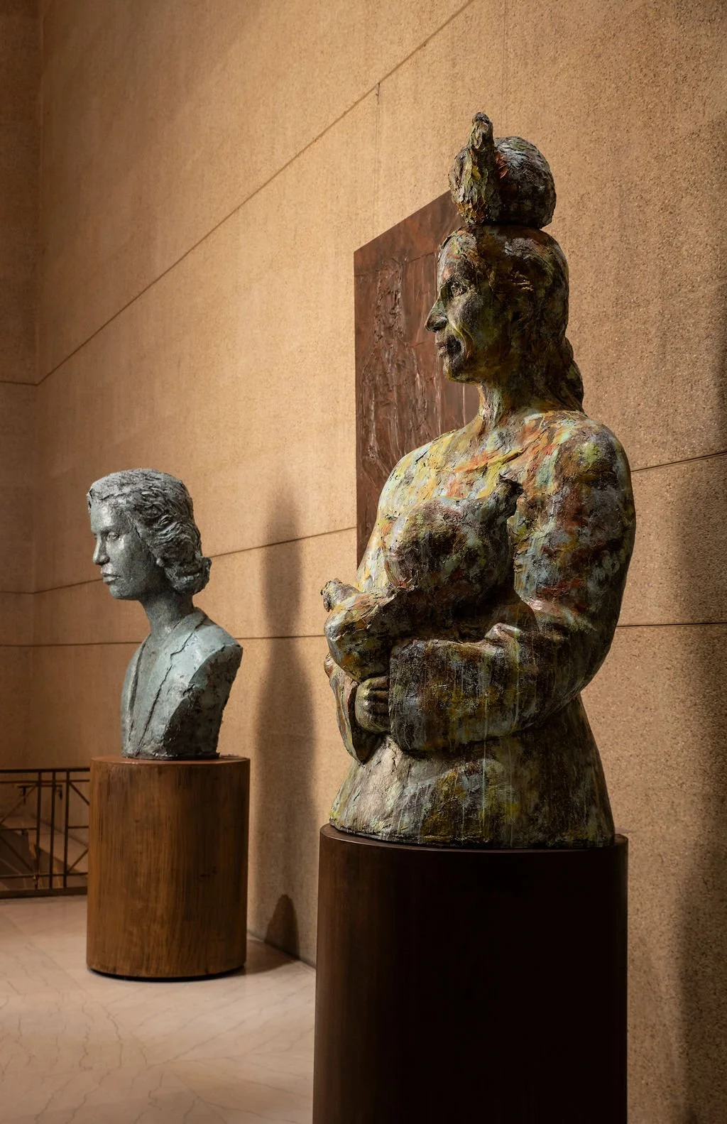 Two bust sculptures on pedestals displayed in an art gallery, one colorful mixed-media and one gray stone, with a textured beige wall background.