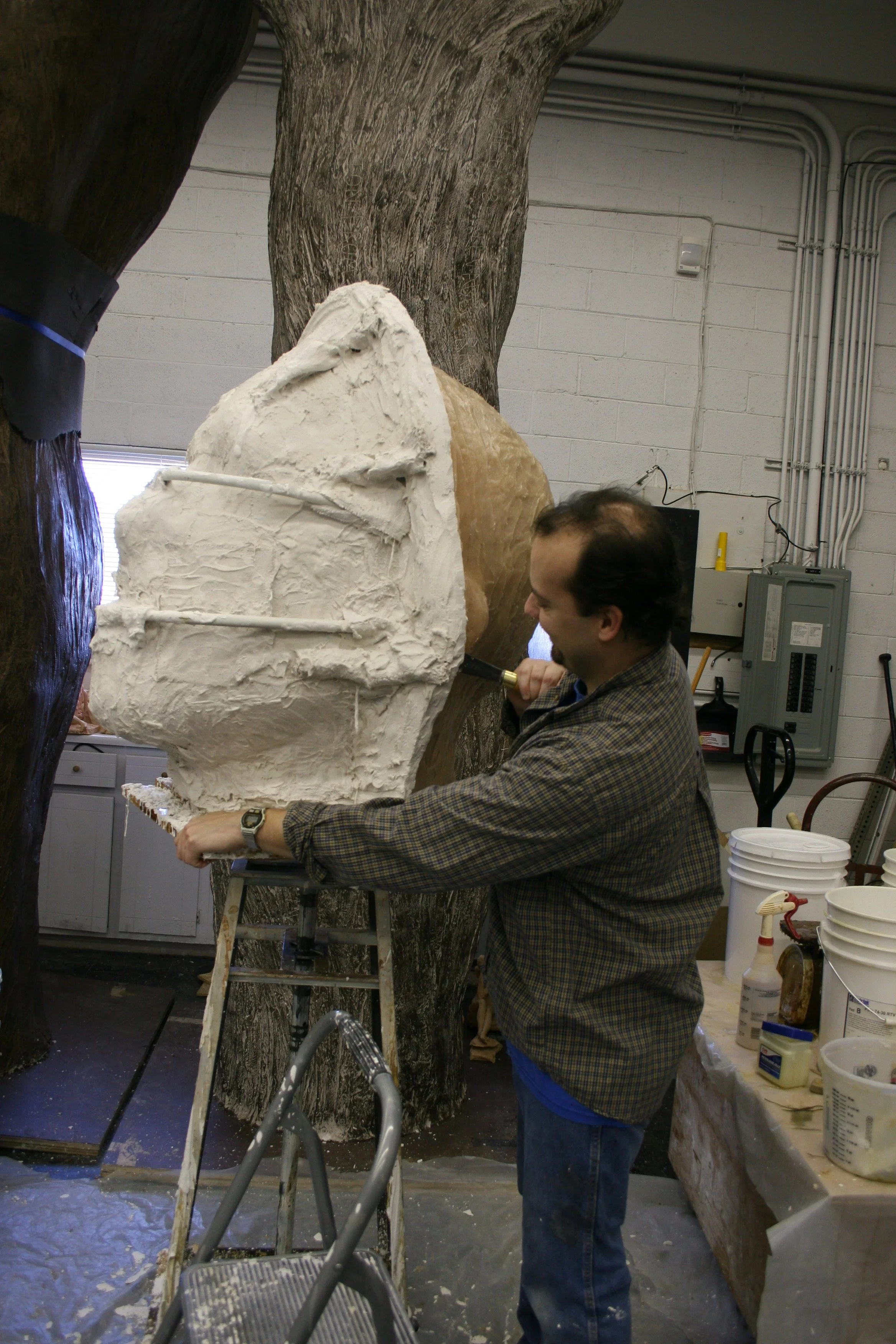 A man with dark hair, wearing a plaid shirt and a blue shirt underneath, is working on a large mother mold. The sculpture is standing on a similar pedestal inside a studio with various tools, buckets, and electrical panels visible in the background.