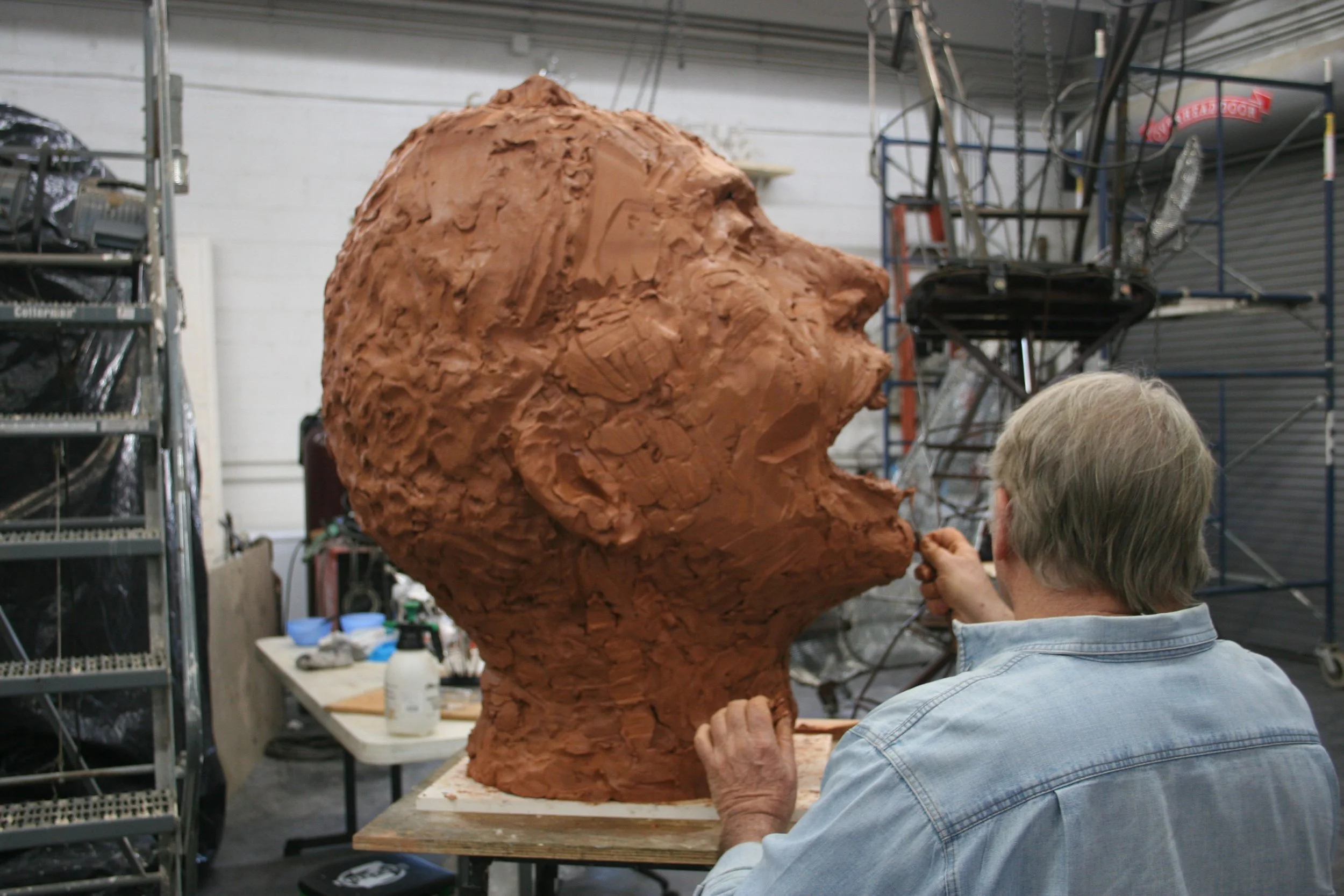 An artist working on a large sculpture Pete Seeger of a person's head in an art studio, with tools and shelves in the background.