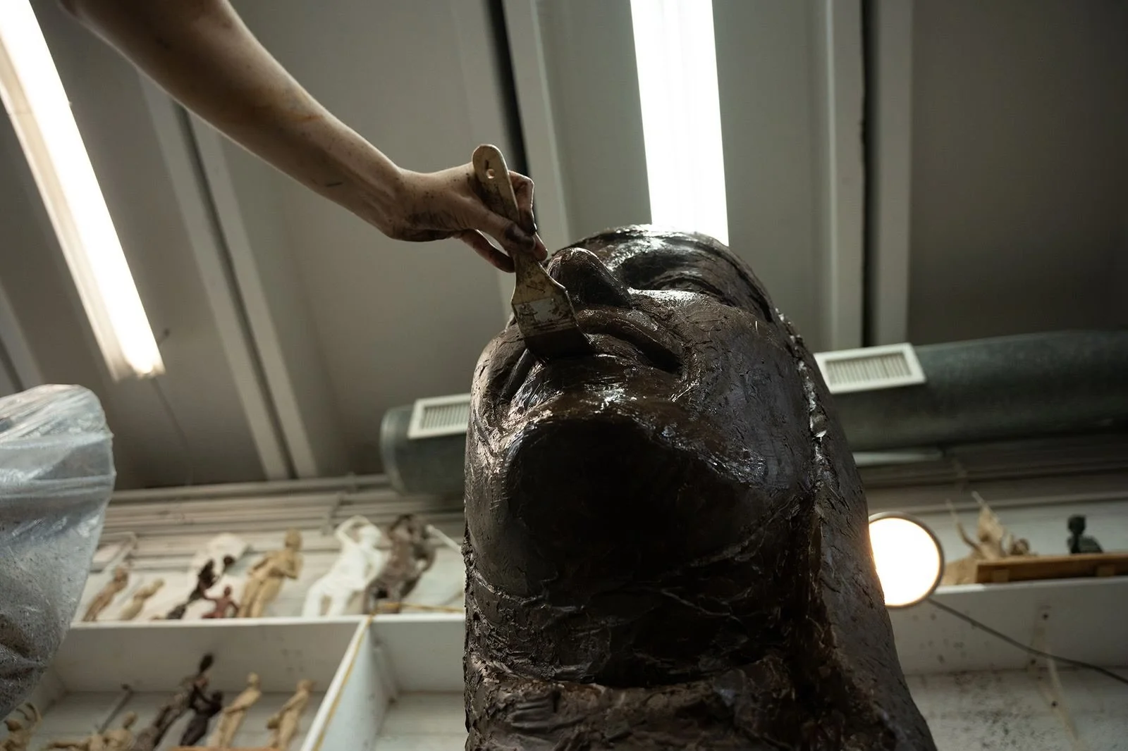 A person carving details into a large, dark sculpture of a human face, with an indoor workshop setting in the background.