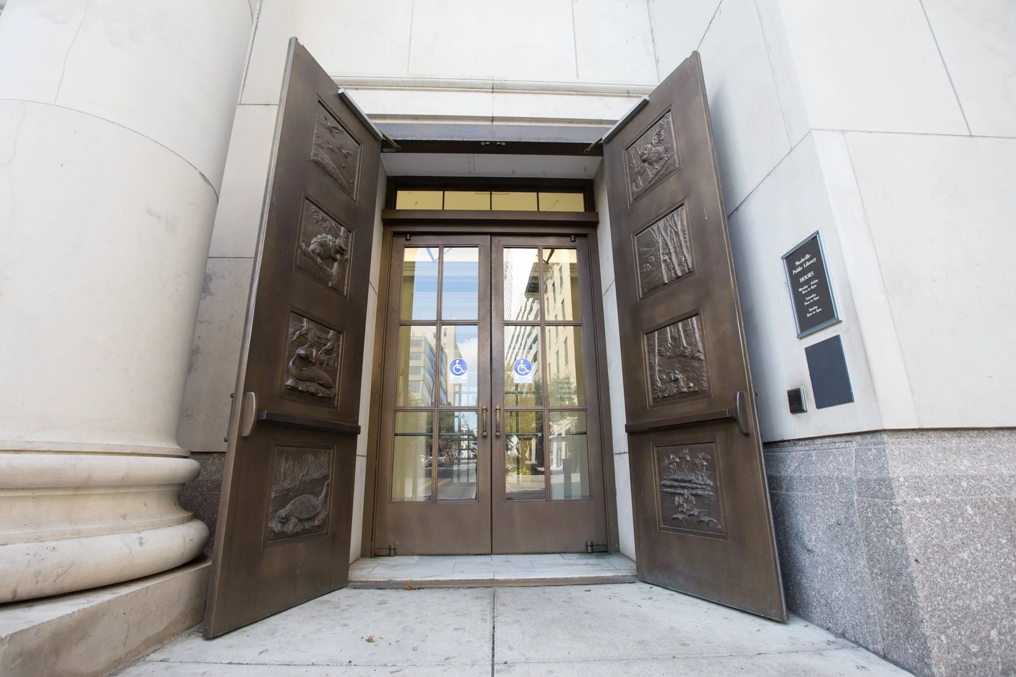 The image shows the entrance of a building with open brown doors. The doors have decorative panels with images, and there are signs indicating accessibility for individuals with disabilities on the glass doors. The building's exterior features large white marble columns and walls.