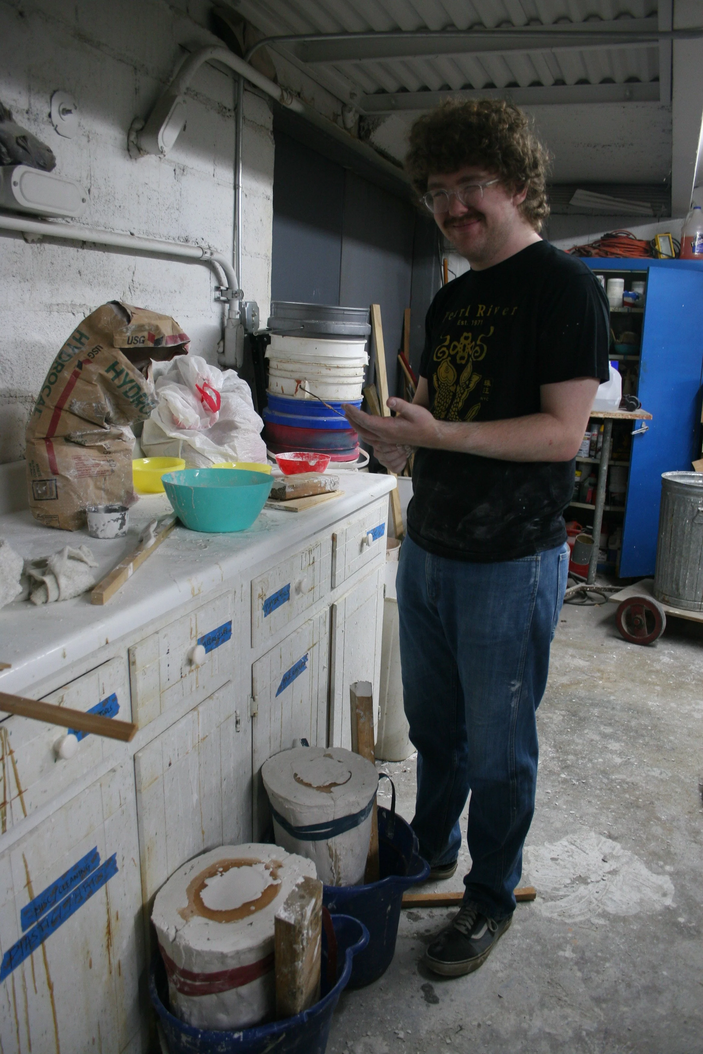 A young man with curly hair, glasses, and a mustache stands in an art or pottery studio next to a counter with bags of clay, bowls, and tools. He is smiling and holding a piece of clay or a sculpted object, wearing a black T-shirt and jeans.