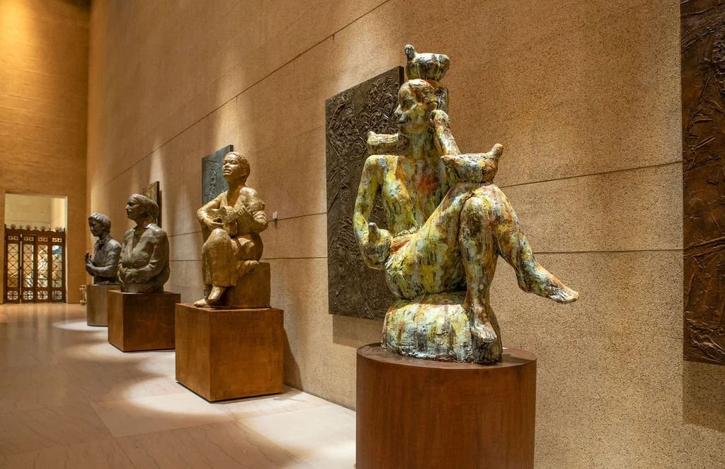 A row of four bronze sculptures of women, each seated on a pedestal and displayed along a beige wall in a museum hallway.