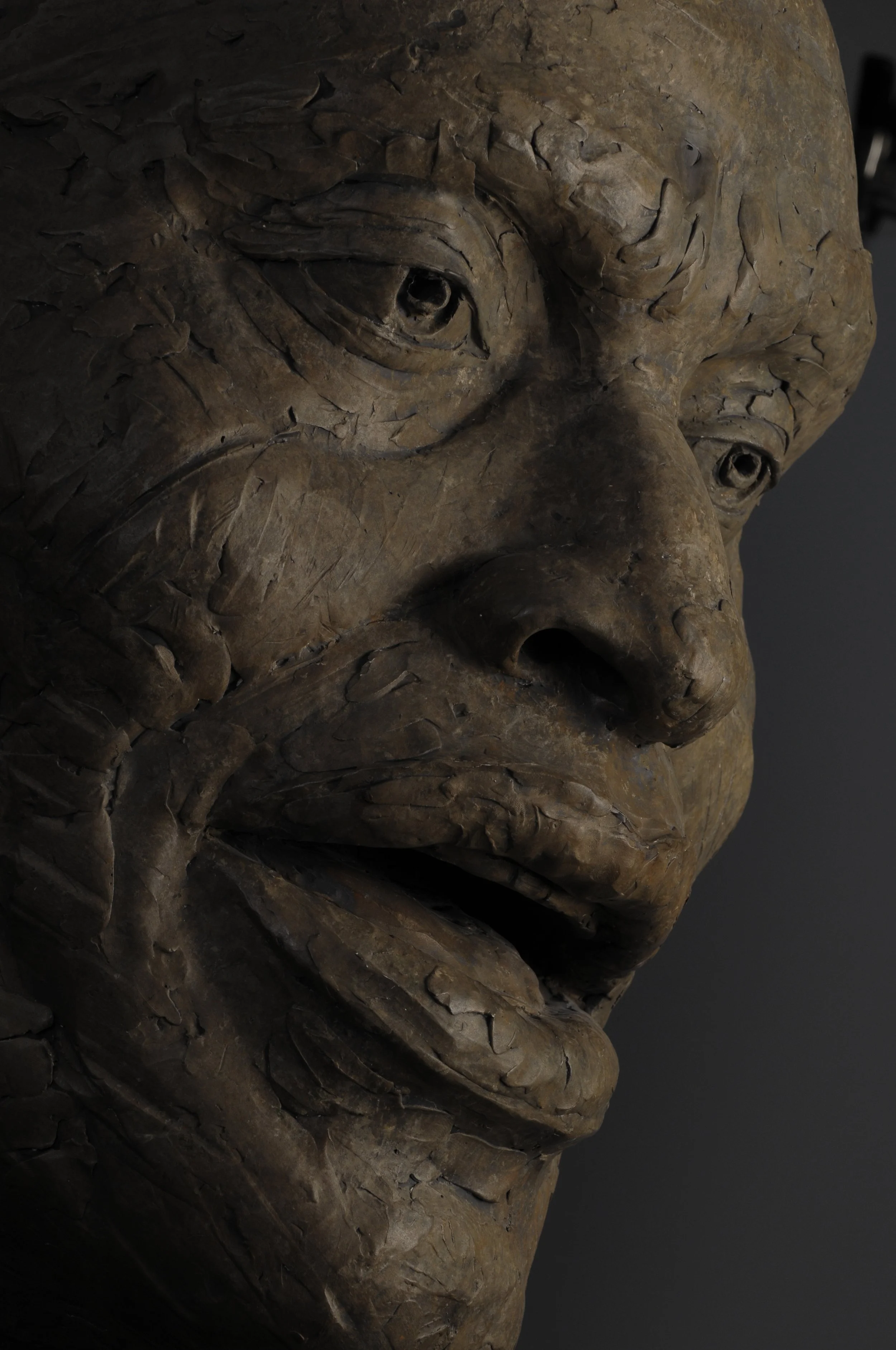 Close-up of a bronze or metal sculpture depicting a human face with detailed wrinkles, textured skin, and expressive eyes.