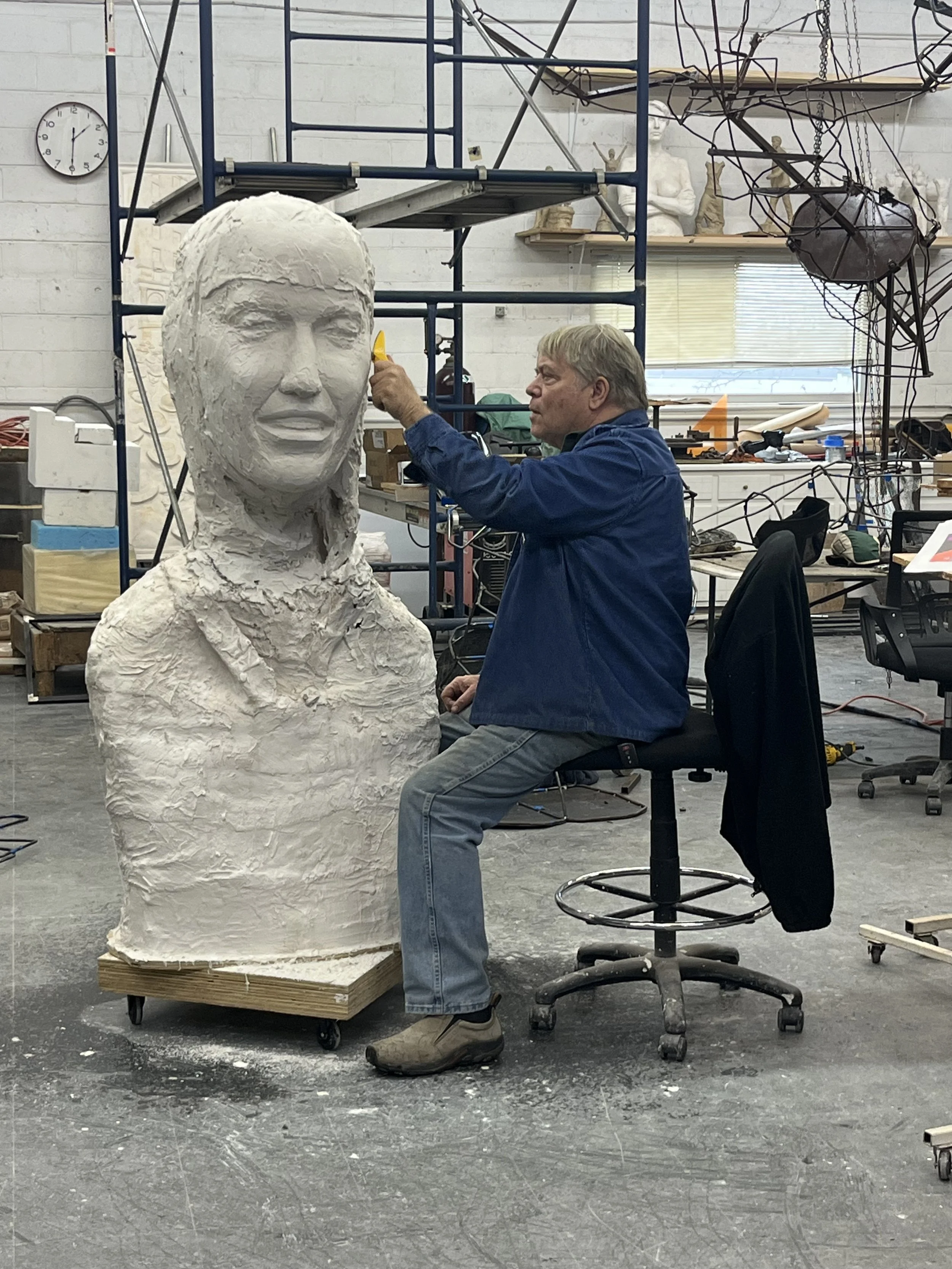 An older man working on a large white sculpture of a woman's head in an art studio.