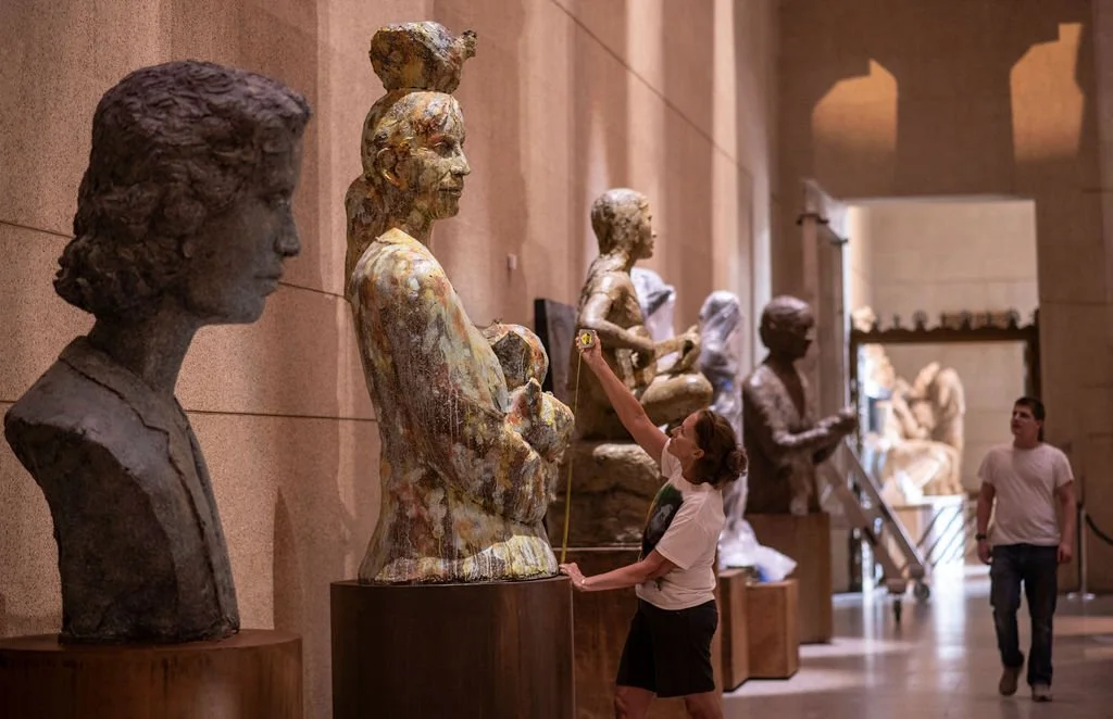 A woman measuring a bust sculpture of a woman at the museum, with other sculptures displayed along the corridor.