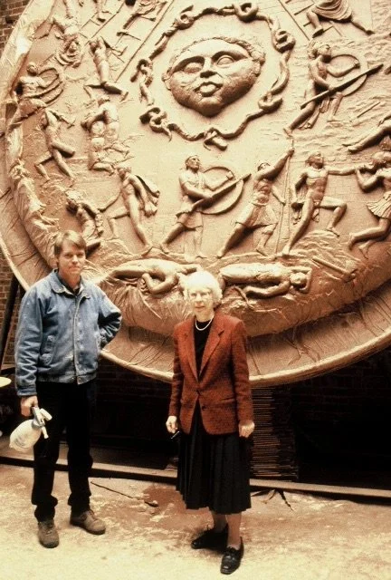 A large ornate wall sculpture with a central face surrounded by figures  of Athenas Shield. Athena Parthenos in Parthenon Museum Nashville. . Two people, a man and a woman, stand in front of the sculpture.