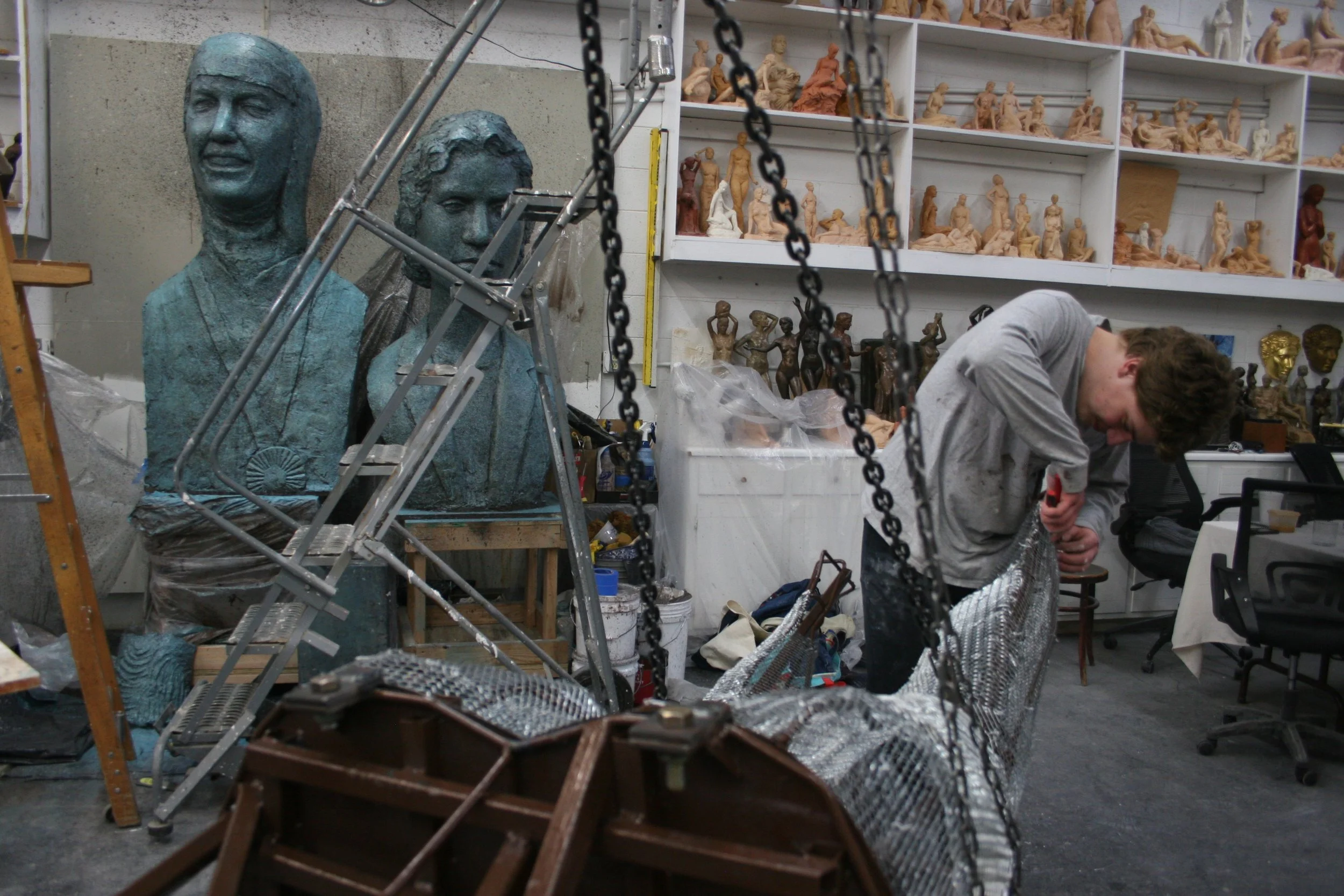 A person working in a sculpture studio with numerous clay sculptures and busts on shelves and a partially finished large bust sculpture in the foreground.