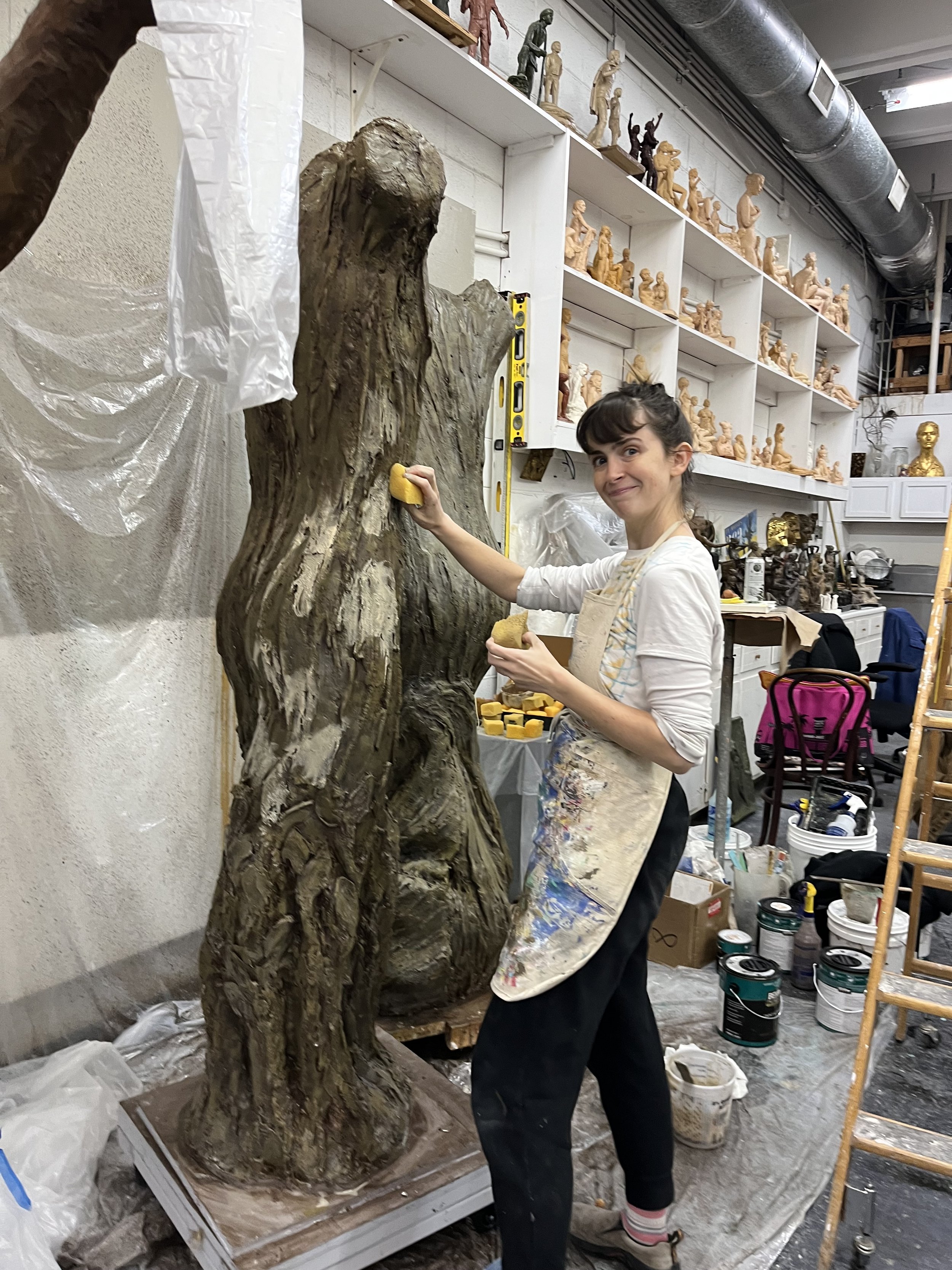 A woman sculptor working on a large wooden sculpture in her art studio, surrounded by shelves filled with small figurines and art supplies.