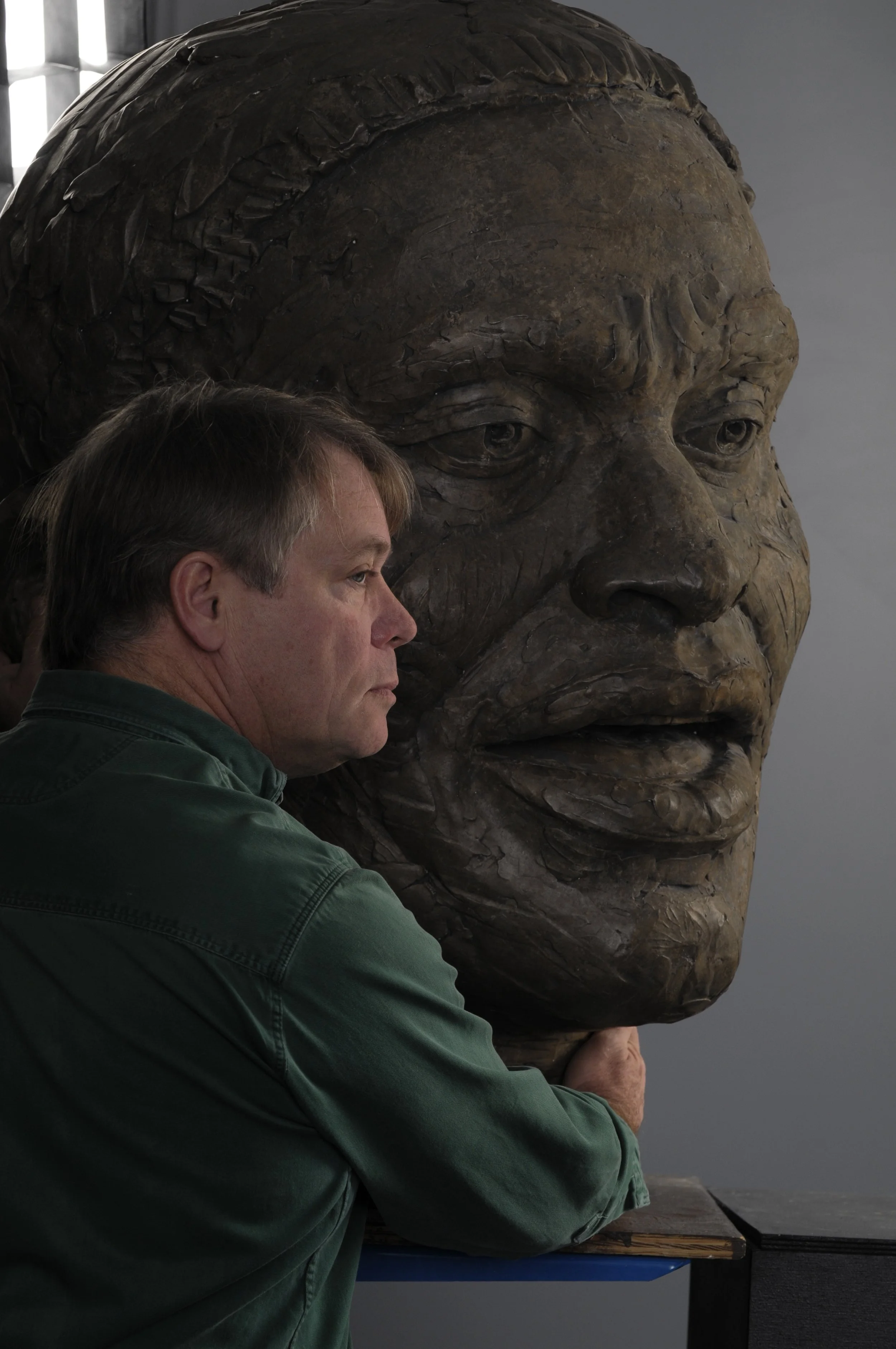 A man with light brown hair and a green shirt touching a large bronze bust of a man with a stern expression.  Cultural Heroes