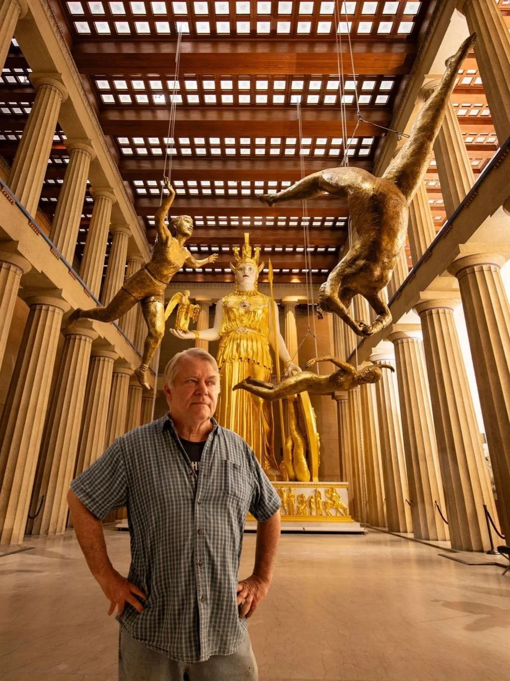 A man standing in a grand hall with classical architecture featuring tall columns and a wooden ceiling, with a large golden statue of a goddess and two large suspended sculptures of humans in dynamic poses above him at the Parthenon Museum