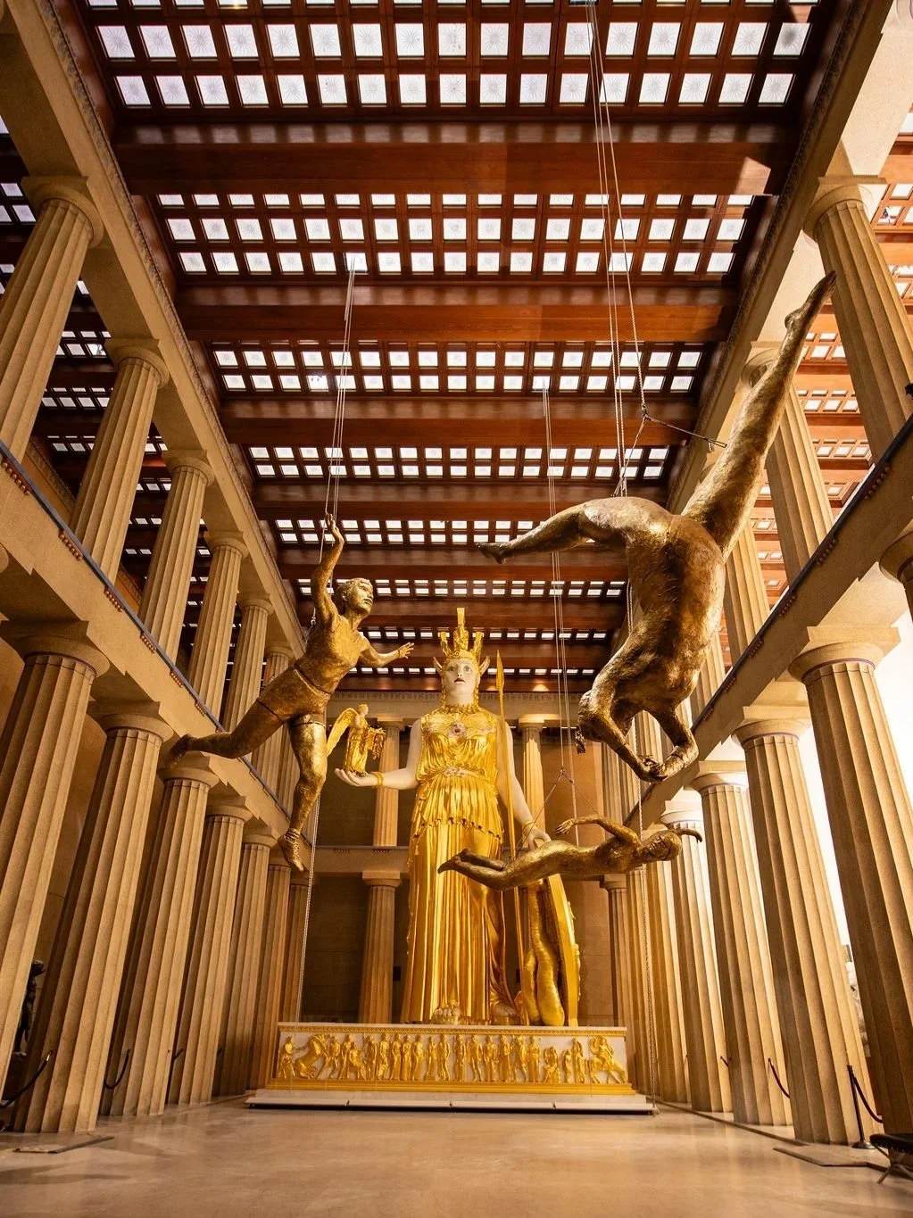 A grand hall with tall columns and a wooden ceiling featuring a large gold and white sculpture of a woman wearing a crown. Surrounding her are four suspended, dynamic gold figures resembling humans in motion. Parthenon Museum
