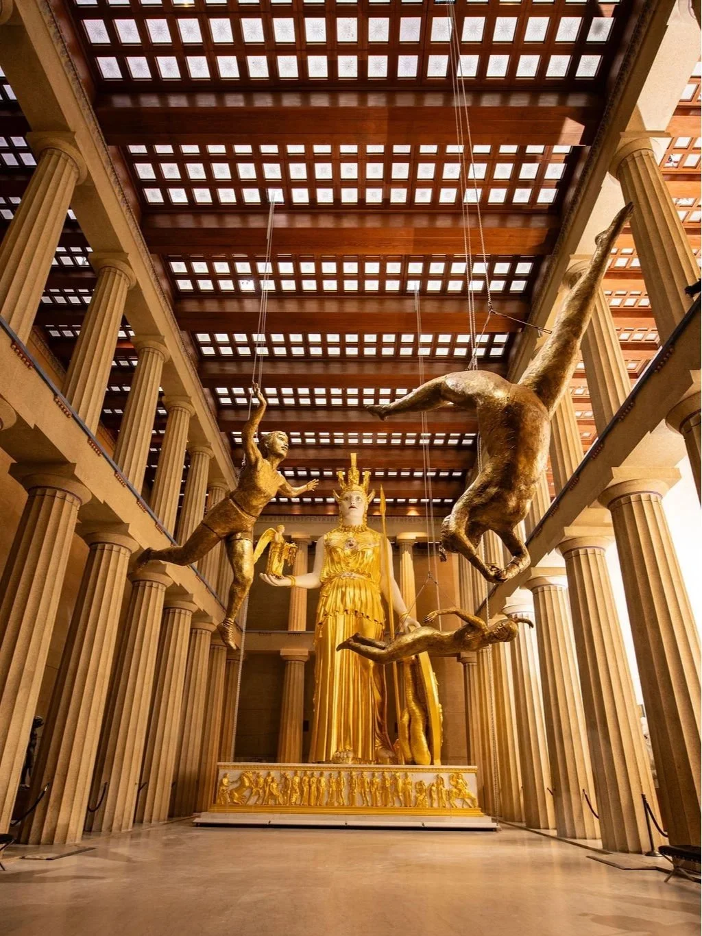 A large golden statue of a woman in a robe, surrounded by columns, with a collection of smaller figures along the base. Above, four large gold sculptures of figures are suspended from the ceiling, appearing to dance or leap in the air, with a wooden 