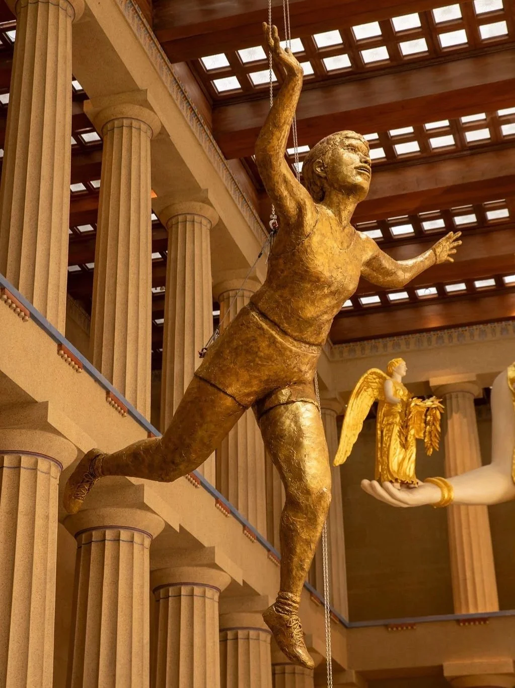 Golden sculpture of a woman with short hair, running with a joyful expression, in a classical building with large columns and a wooden ceiling. At the Parthenon Museum.