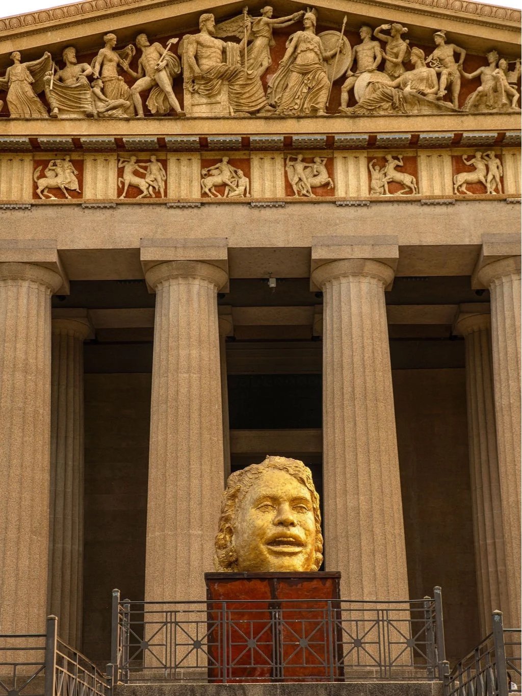 Large sculpture of a smiling child's head in front of a classical building with tall columns and detailed sculptures. At the Parthenon Museum