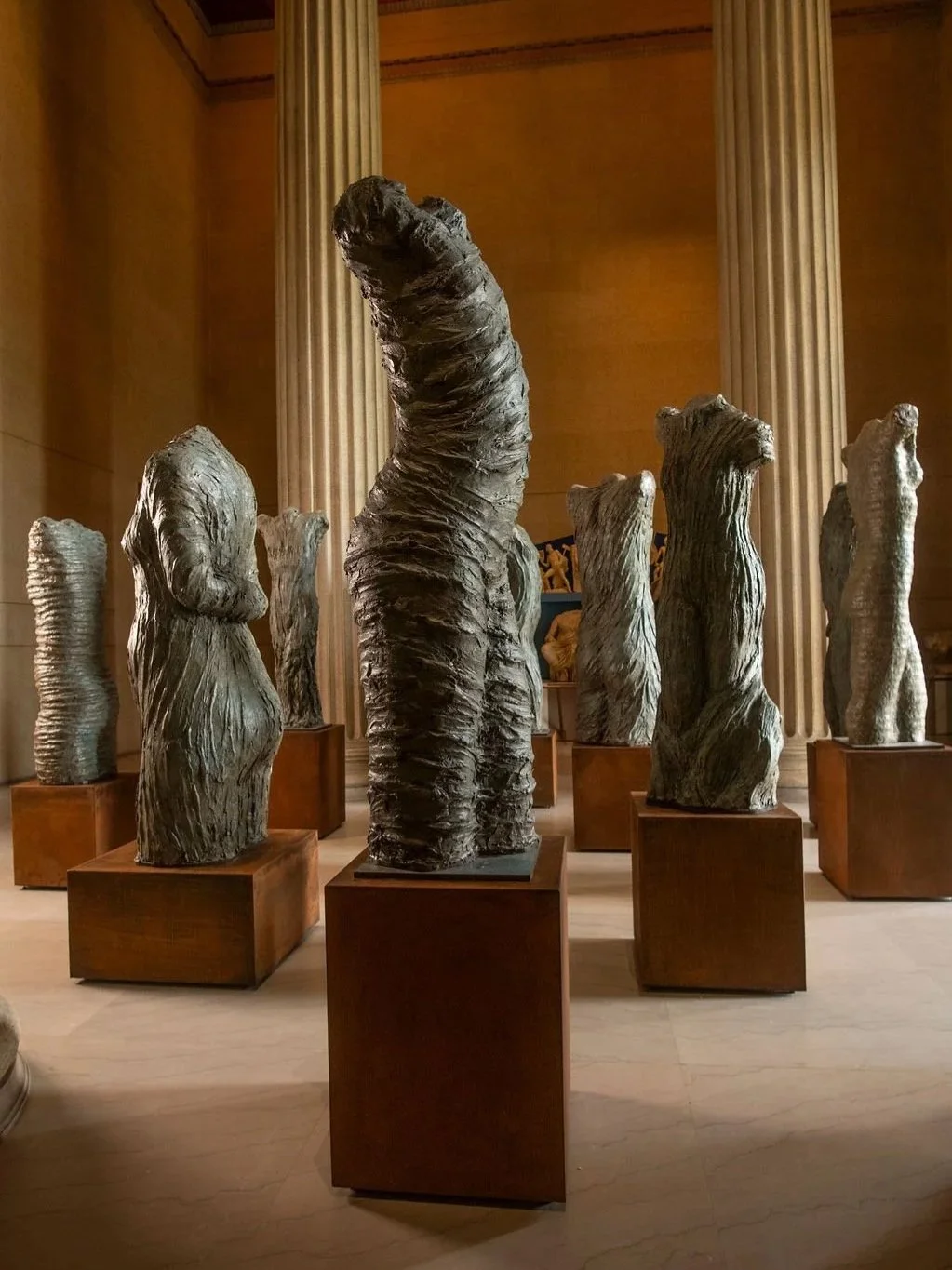 Several large, abstract, textured sculptures resembling human torsos displayed on metal pedestals in a museum gallery with tall columns and warm lighting. At the Parthenon Museum.