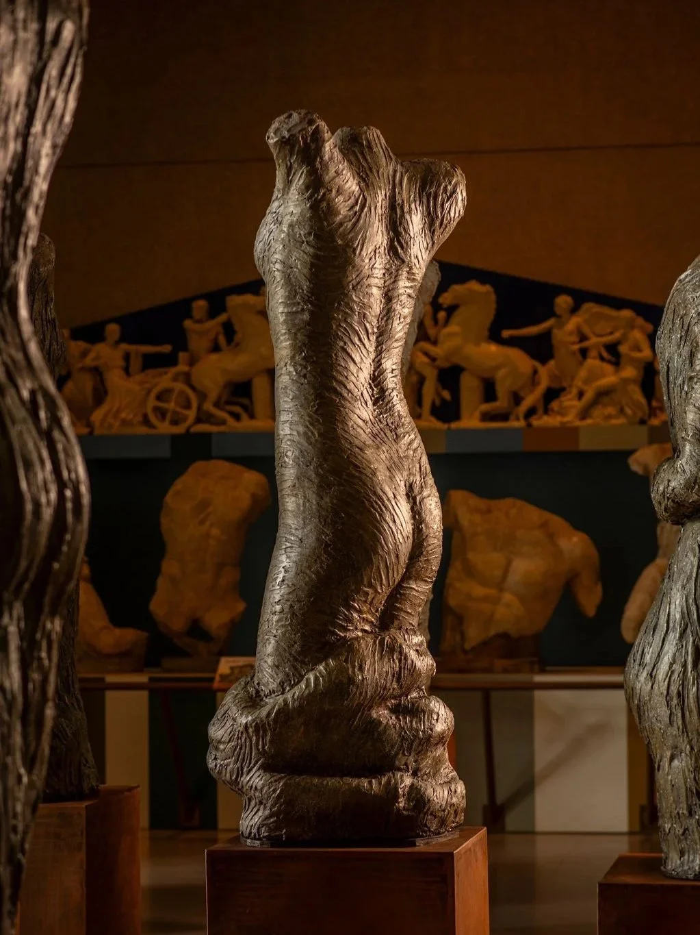 Plaster sculpture of a female torso displayed on a metal stand in the Parthenon museum, with other sculptures and artworks in the background.
