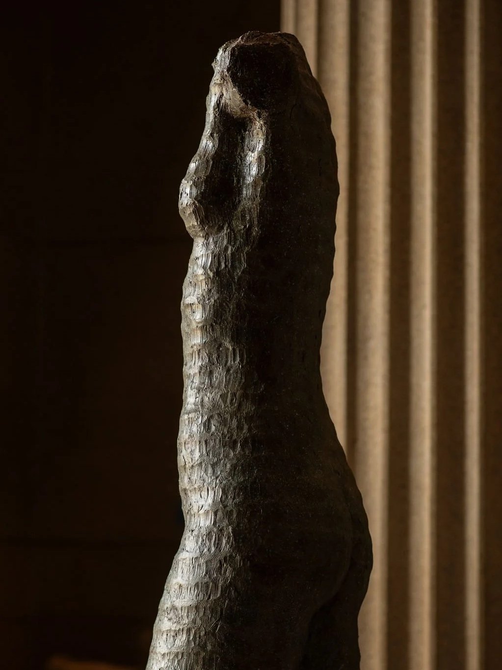 A dark, textured sculpture resembling a woman with a curvy figure, standing indoors with column in the background. At the Parthenon Museum.