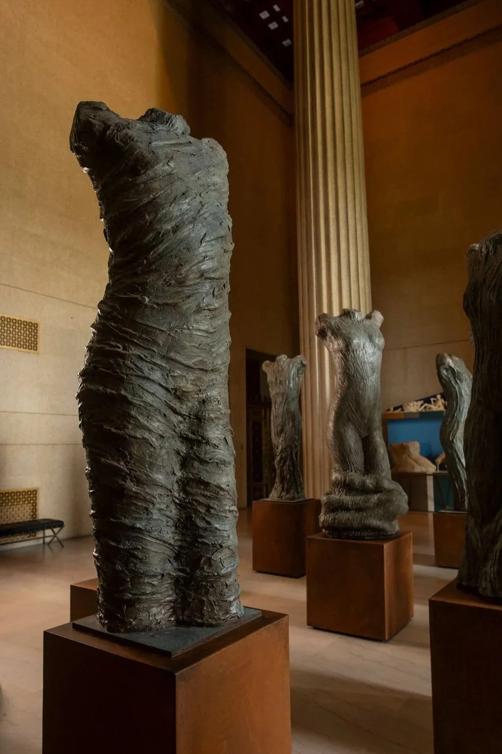 Indoor art gallery displaying tall, abstract, textured sculptures of human torsos, placed on metal pedestals with a large classical column in the background.