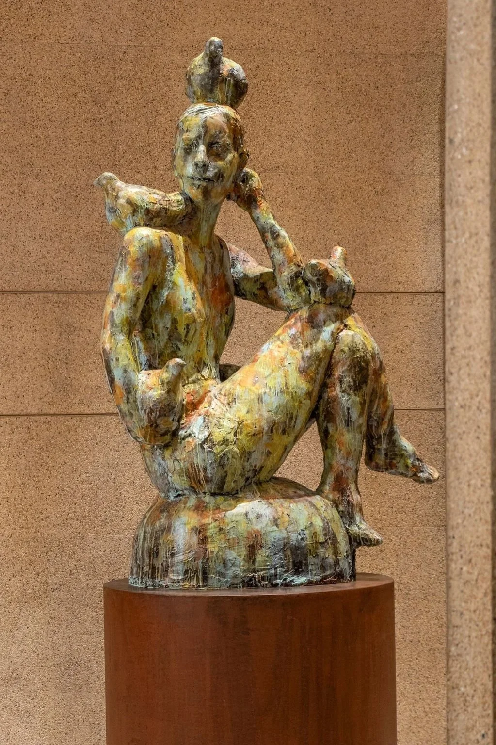 Colorful abstract sculpture of a seated woman with her knees up, holding her chin with her hand, and a bird on her shoulder, on a steel pedestal against a brown wall.