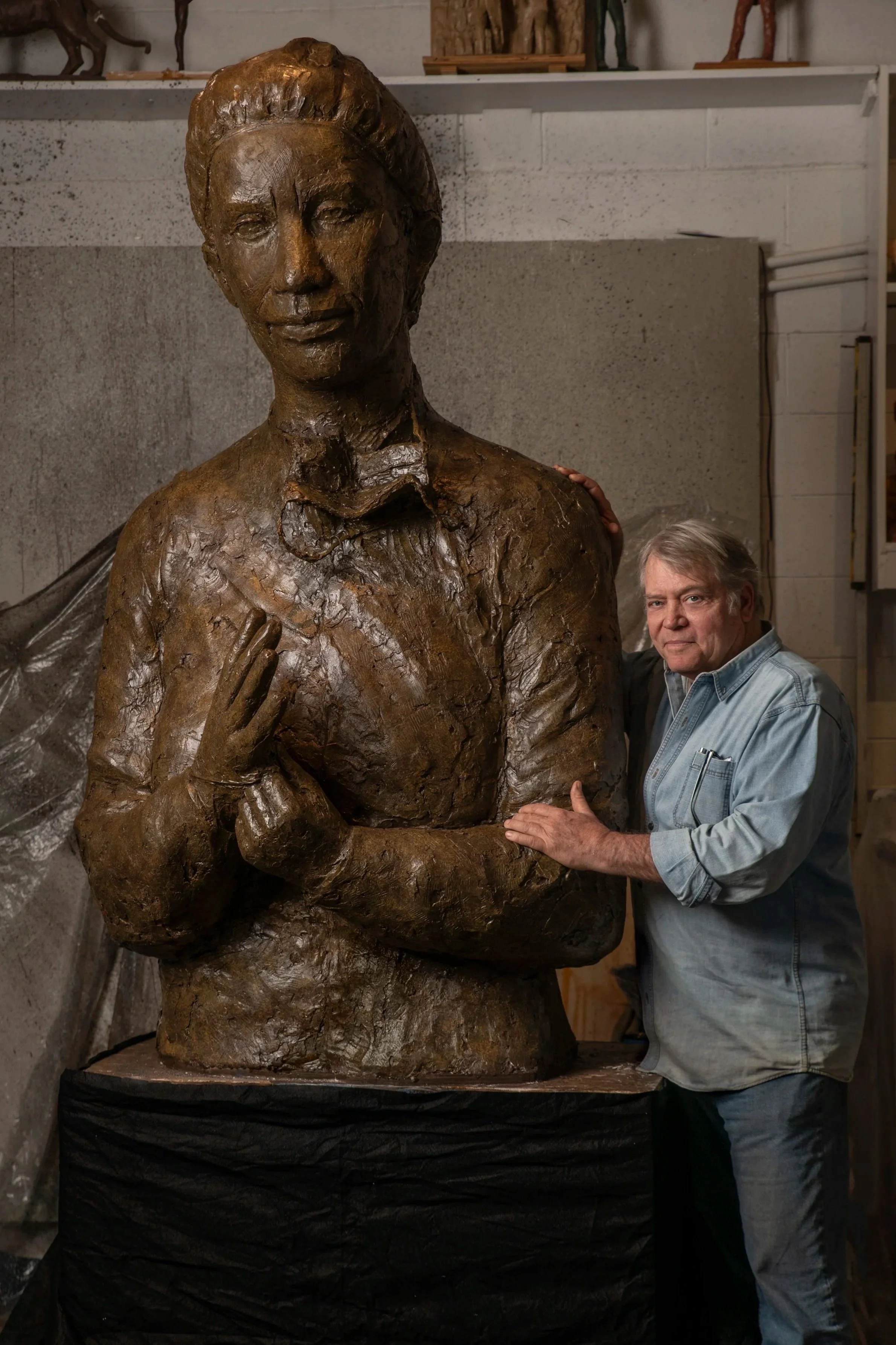A man is standing beside a large bronze sculpture of a woman, in an artist's studio or workshop. The sculpture depicts a woman with short hair, a bun, and a calm expression, with detailed features and clothing. The man appears to be the artist, inspe