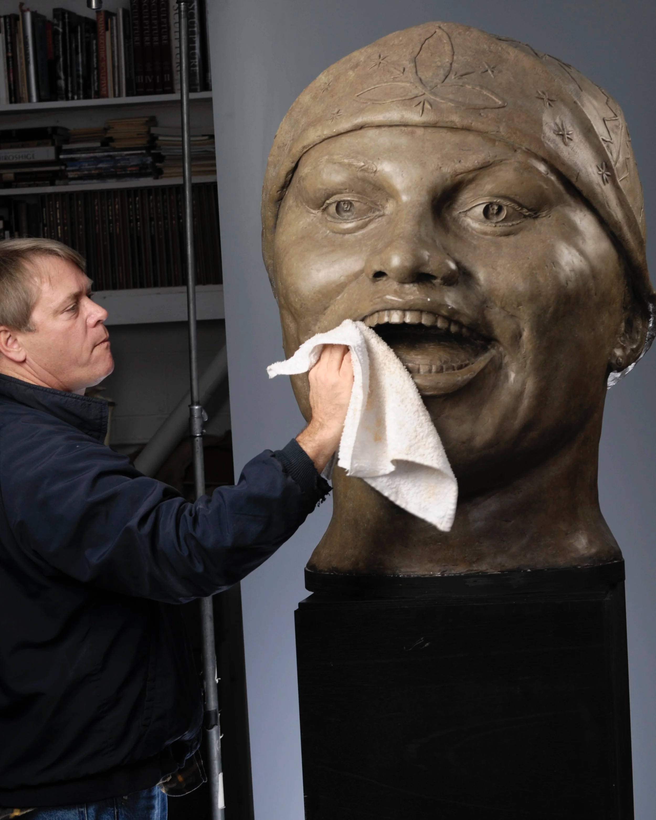 A man polishing a large bronze sculpture of a woman’s face with a white cloth. The sculpture has detailed facial features and is wearing a headscarf with carved symbols. In the background, there is a bookshelf filled with books.  Cultural Heroes