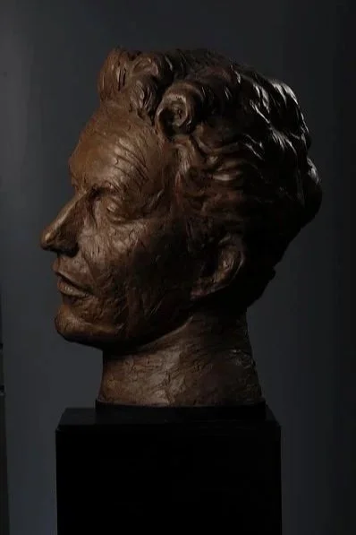Bronze bust sculpture of a Woody Guthry’s head with curly hair on a black pedestal against a dark background.  Cultural Heroes