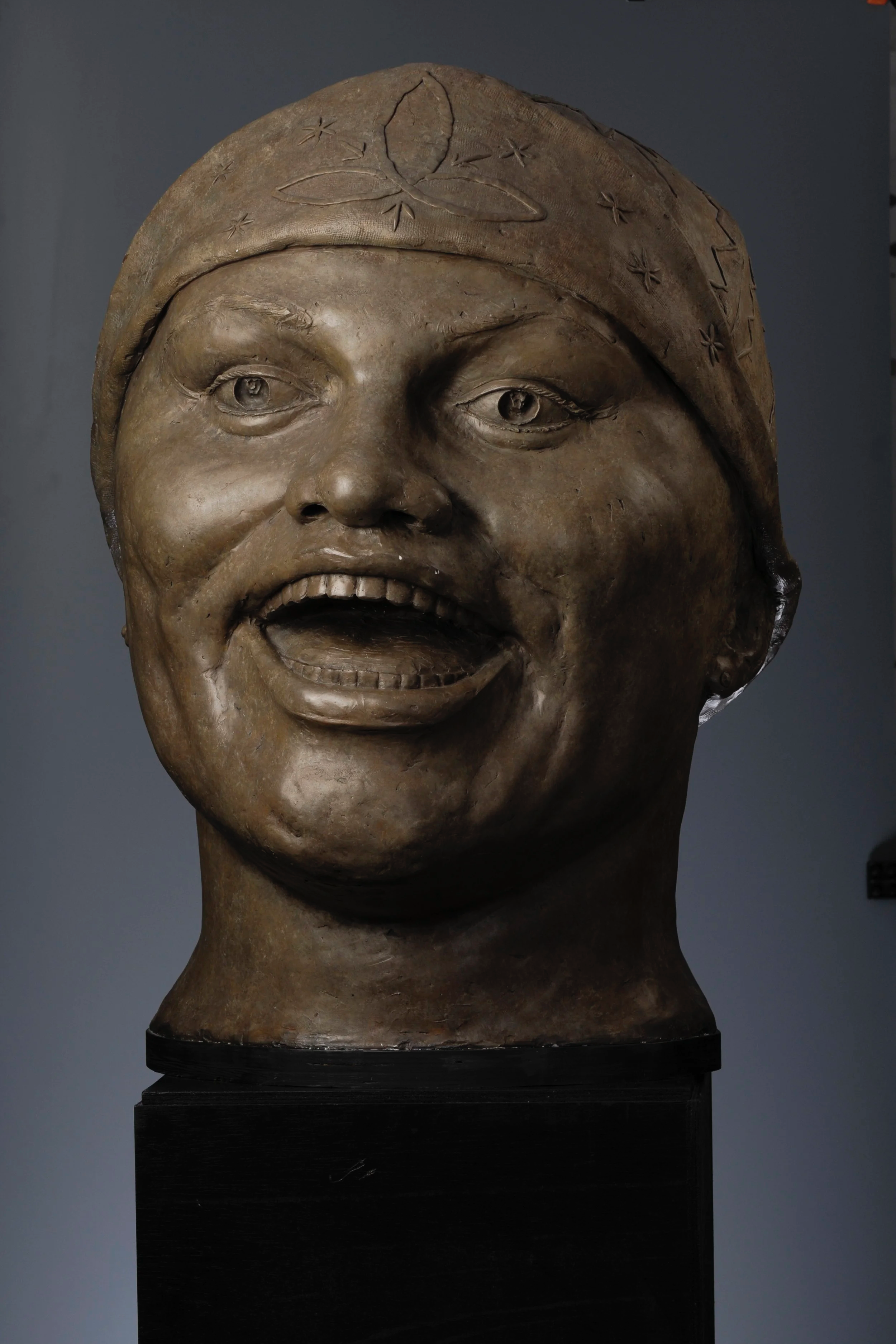 A bronze sculpture of a smiling woman's head with a headscarf decorated with embossed leaves and stars. The sculpture includes detailed facial features and is mounted on a black pedestal.  Cultural Heroes