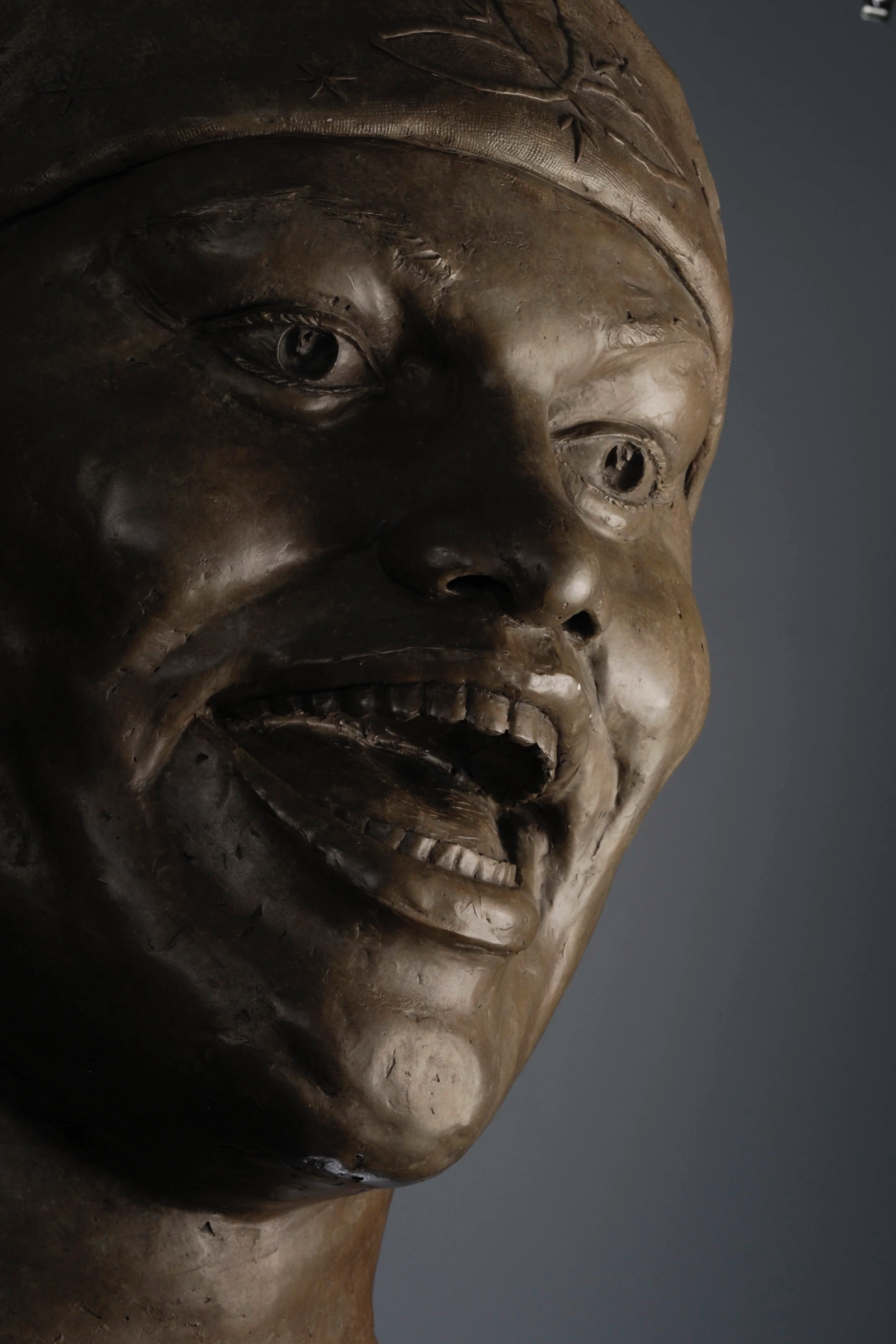 Close-up of a bronze sculpture of a human face, showing detailed features including eyes, nose, mouth, and a head covering or hair. The sculpture has a somber expression and appears to be part of a larger artwork.  Cultural Heroes