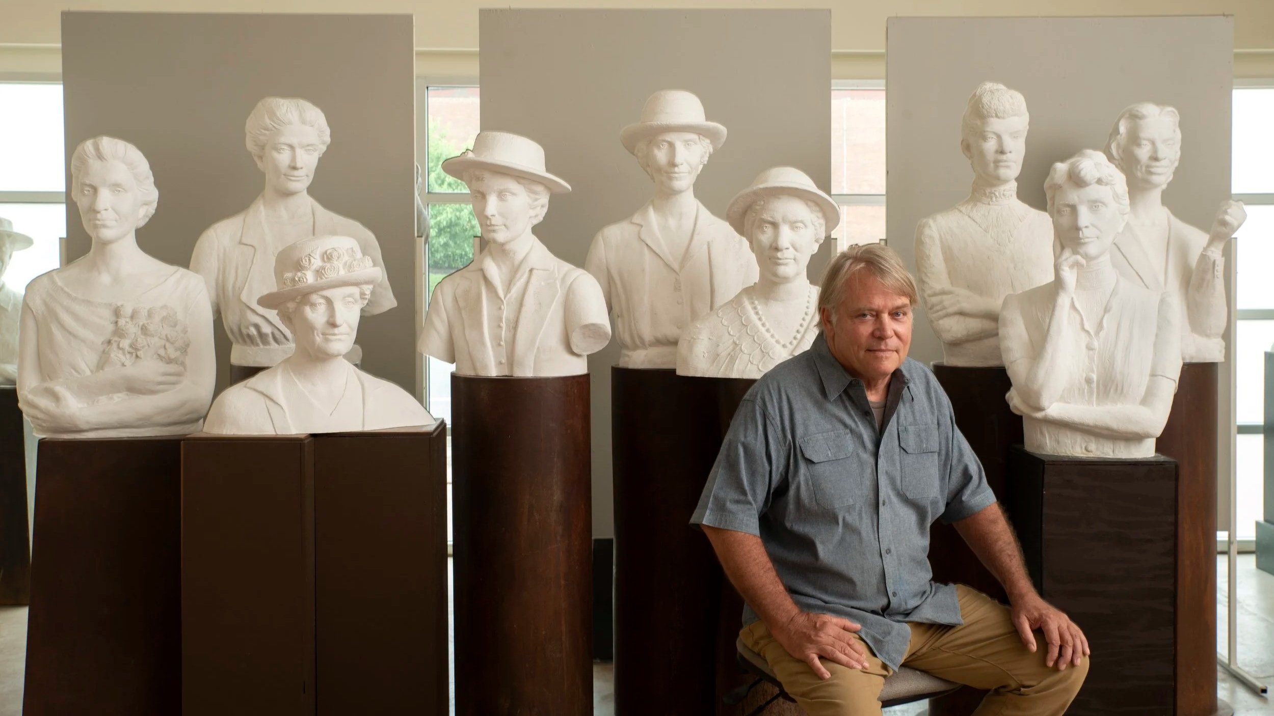 An older man Alan LeQuire sitting in front of a display of white bust sculptures of women in a museum or gallery.