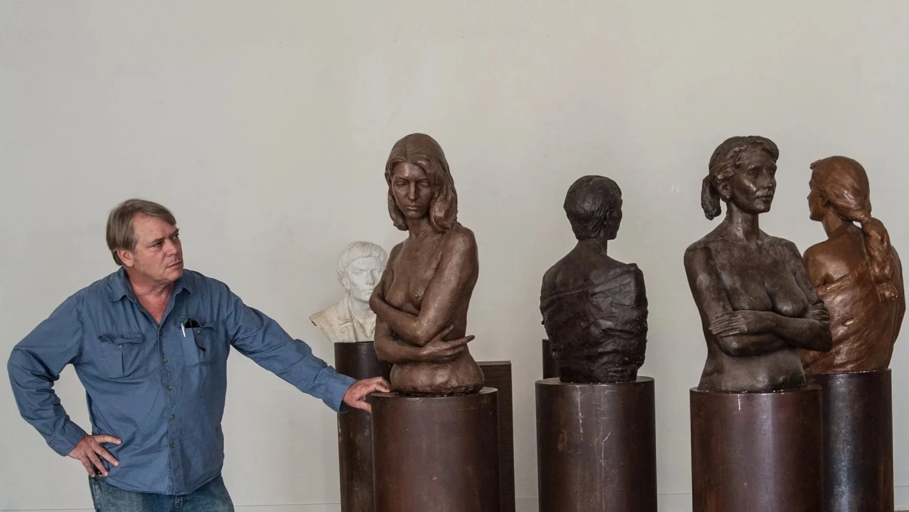 Alan Lequire in a blue shirt looking at a row of bronze bust sculptures of women in an art gallery.