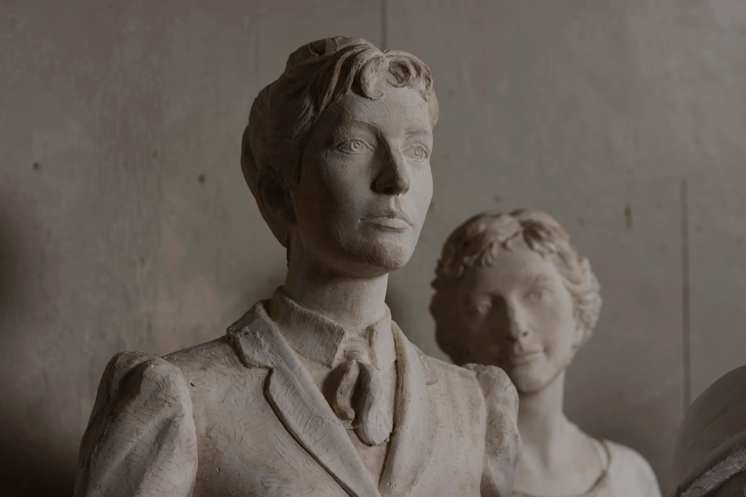 Close-up of classical plaster bust sculptures, focusing on a female figure with detailed facial features and hairstyle, with two other sculptures partially visible behind her. The background is plain and muted. Woman Suffrage