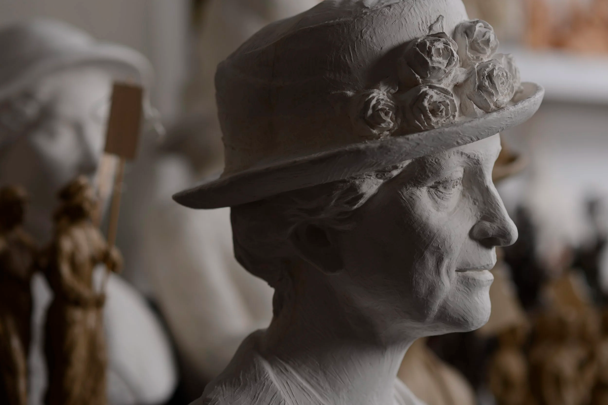 A white sculpture of a woman's head wearing a wide-brimmed hat with roses on it, displayed among other sculptures. Woman Suffrage. Chapman cap