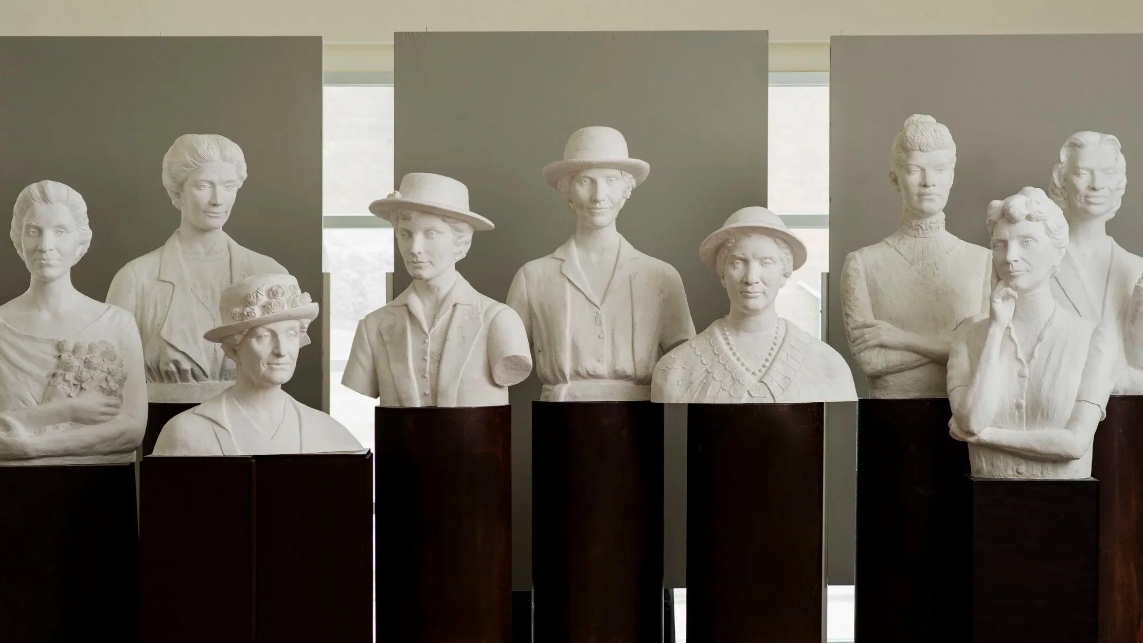 Display of multiple white bust sculptures of women, some wearing hats, with a background of windows and grey walls. Woman Suffrage
