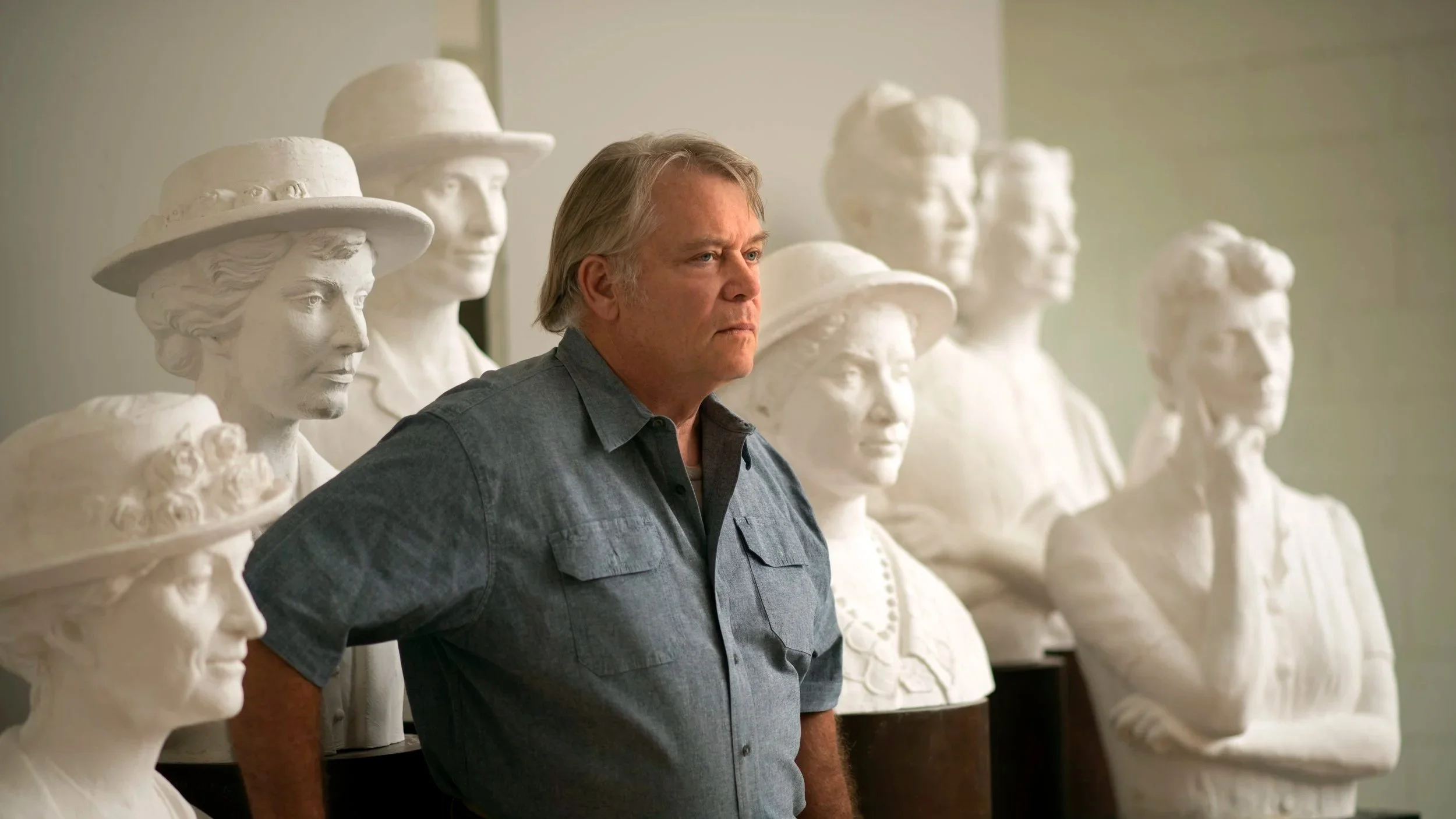 A man stands among a row of white classical bust sculptures of women's heads, some wearing hats, in an indoor setting, possibly a museum or gallery. Womans Suffrage