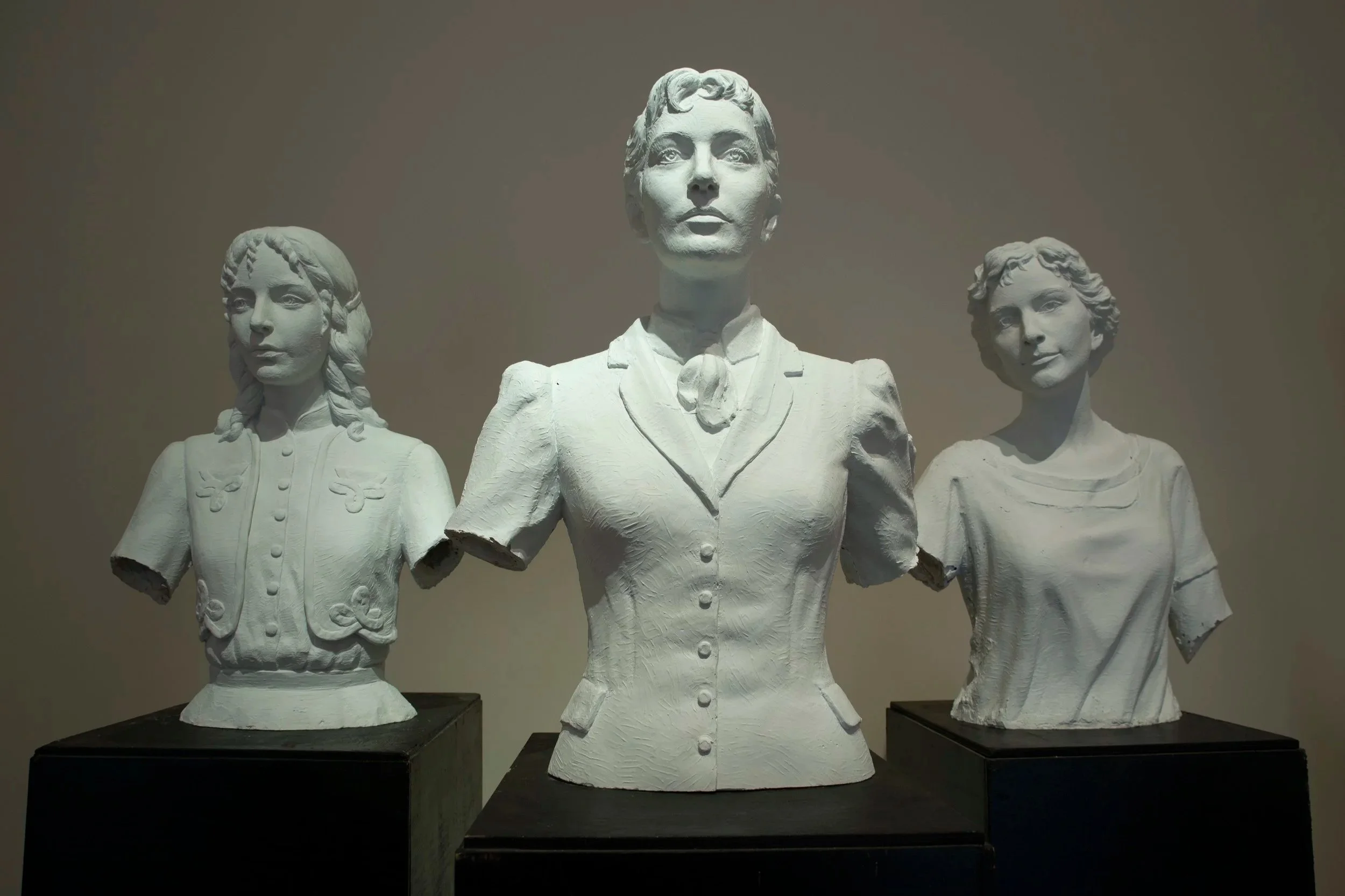 Three white bust sculptures of women displayed on black pedestals, with the central statue being larger and more detailed, set against a plain background. Woman Suffrage