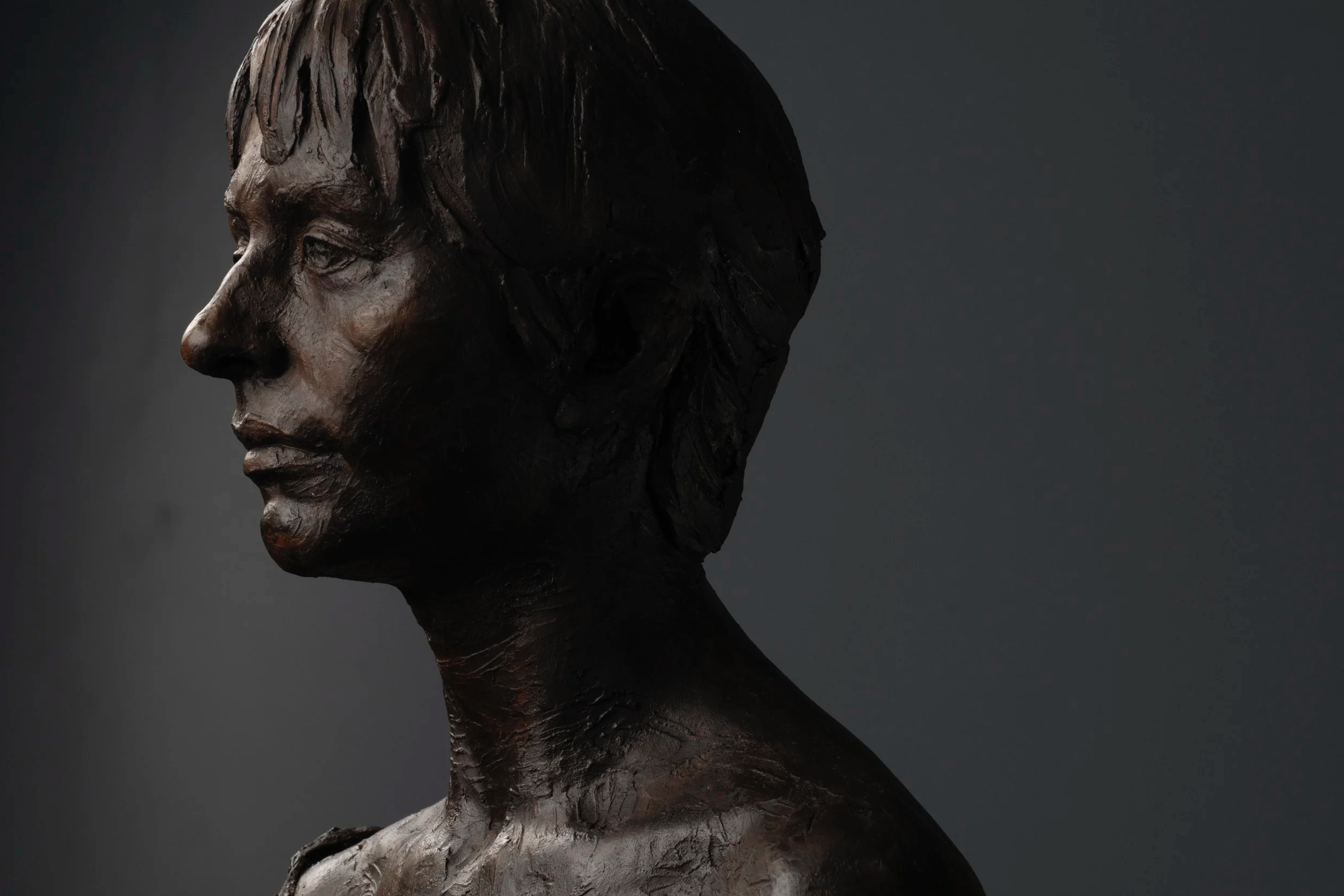 A close-up profile of a bronze or dark metal sculpture of a woman’s face and neck, facing left, with detailed hair and facial features, against a dark background.