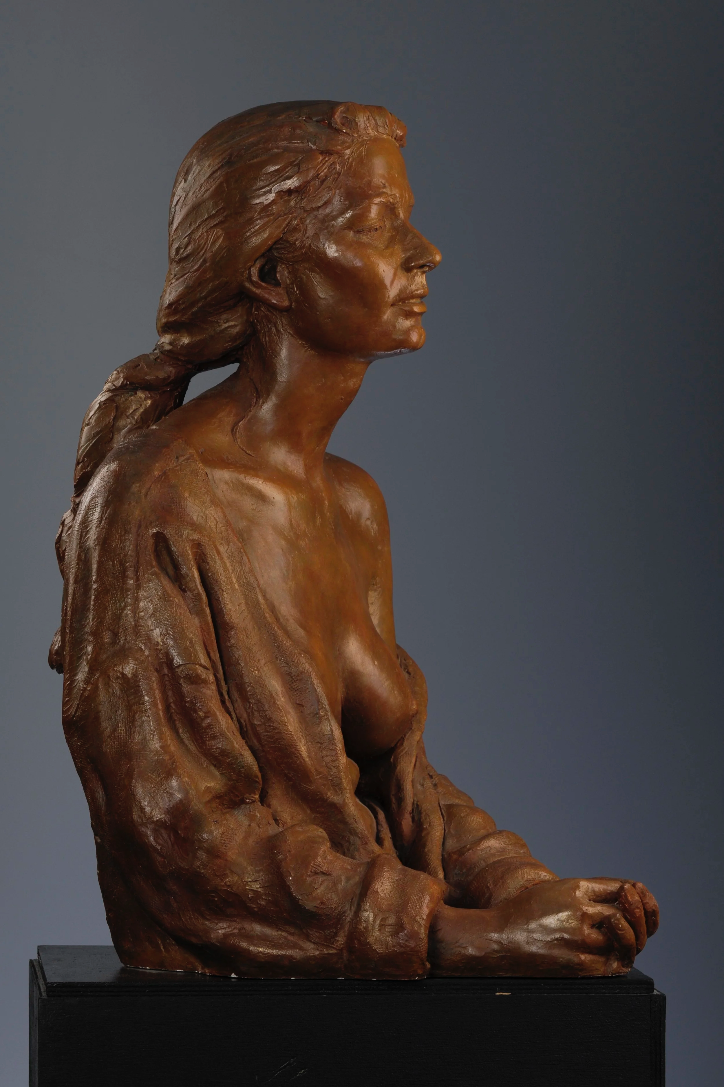 Bronze statue of a woman with closed eyes, sitting with hands clasped, mounted on a black pedestal against a plain gray background.