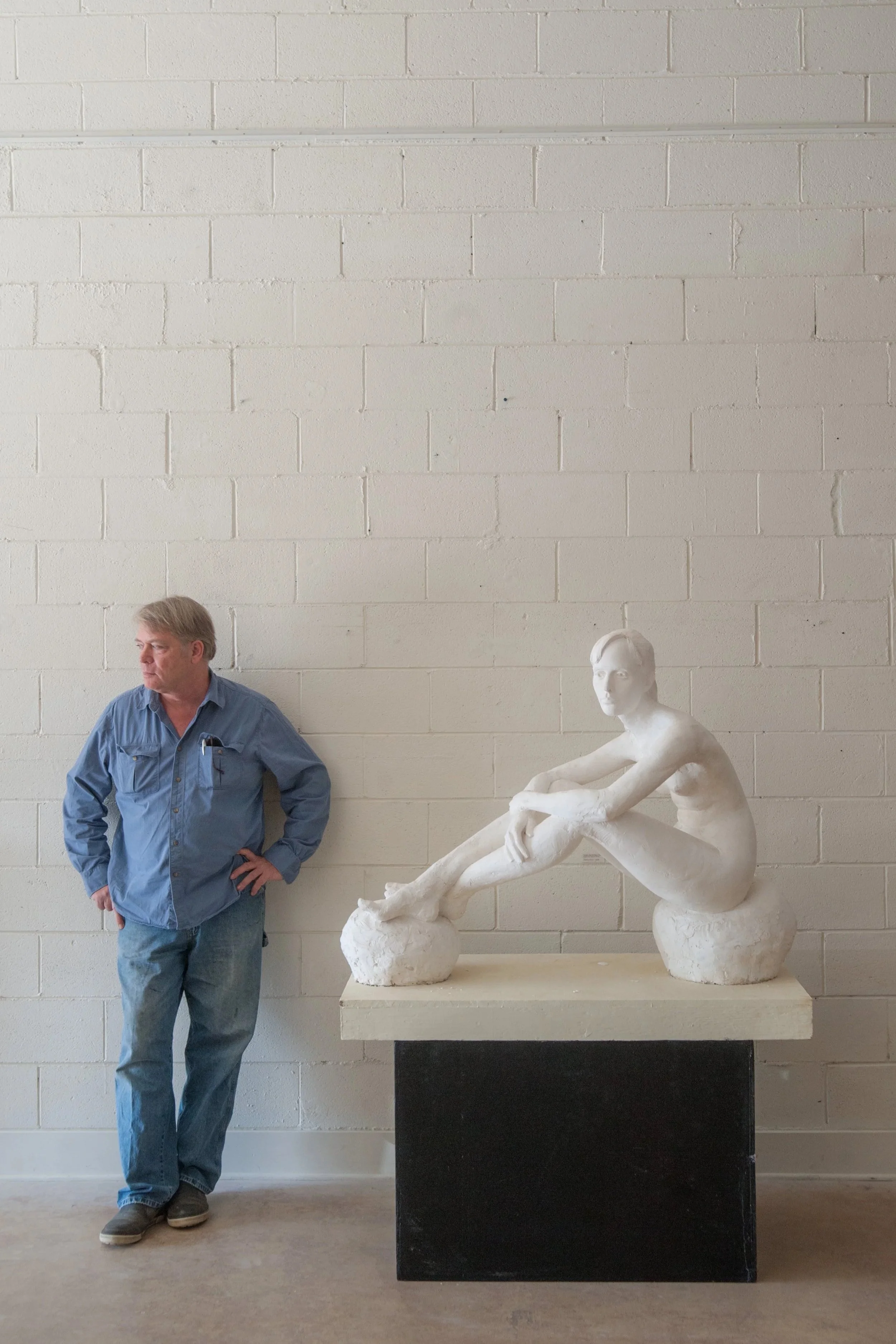 Alan LeQuire with gray hair wearing a blue shirt and jeans standing next to a white seated sculpture of a woman. The sculpture is on a black pedestal against a white brick wall.