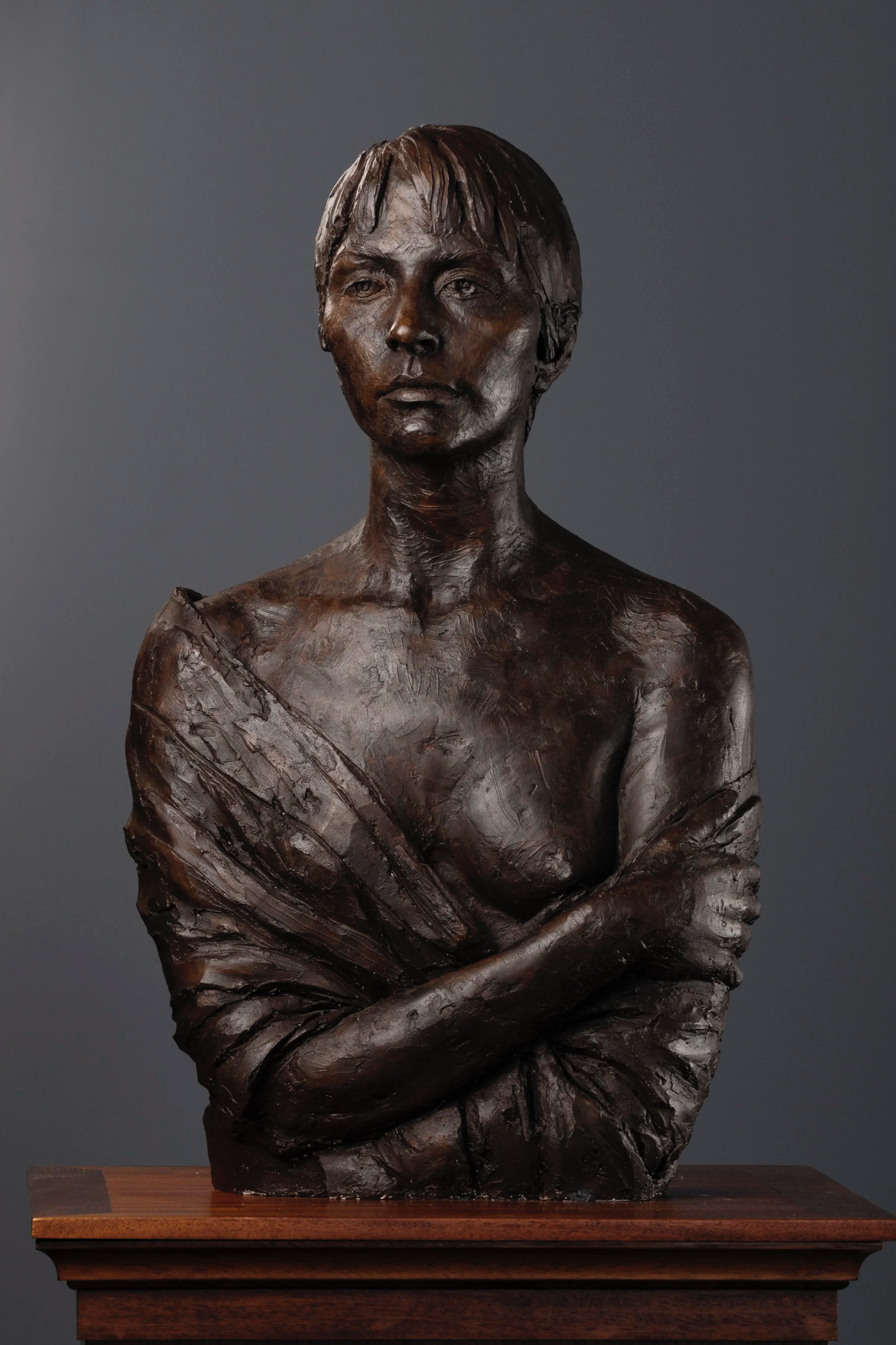 A bronze sculpture of a woman with short hair, her arms crossed, draped in a cloth, mounted on a wooden base.