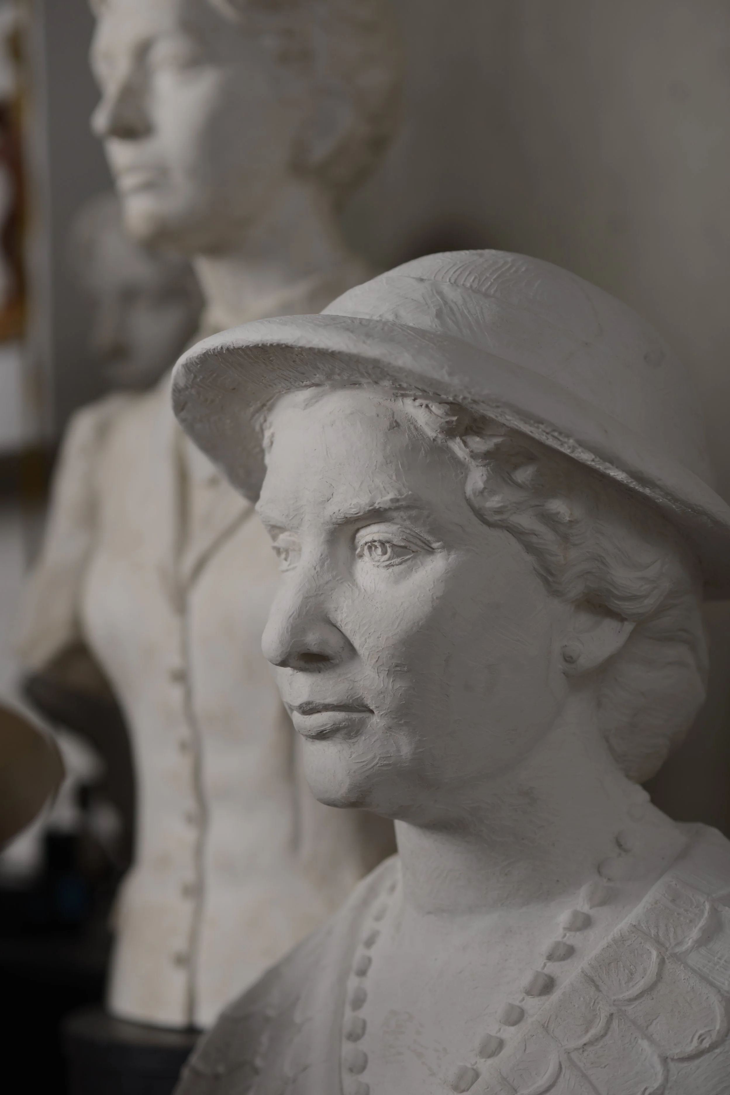 Close-up of a white sculpture bust of a woman's face wearing a large brimmed hat, with other similar busts in the background. Woman Suffrage