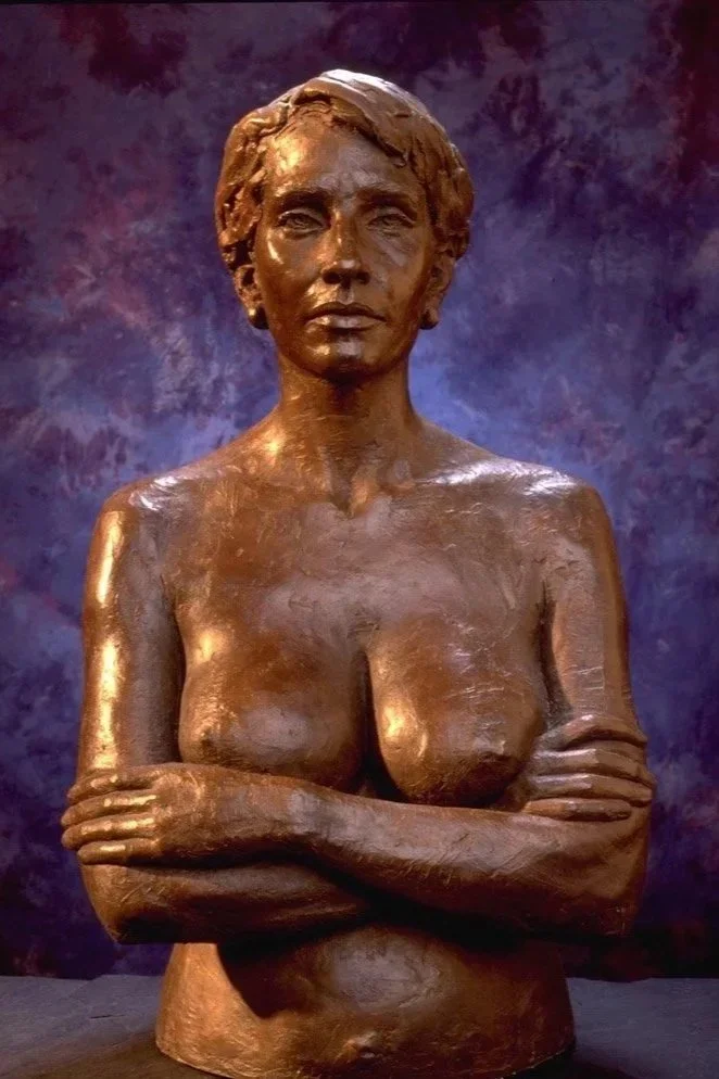 Bronze sculpture of a topless woman with arms crossed, looking forward, with short hair, set against a dark, textured background.