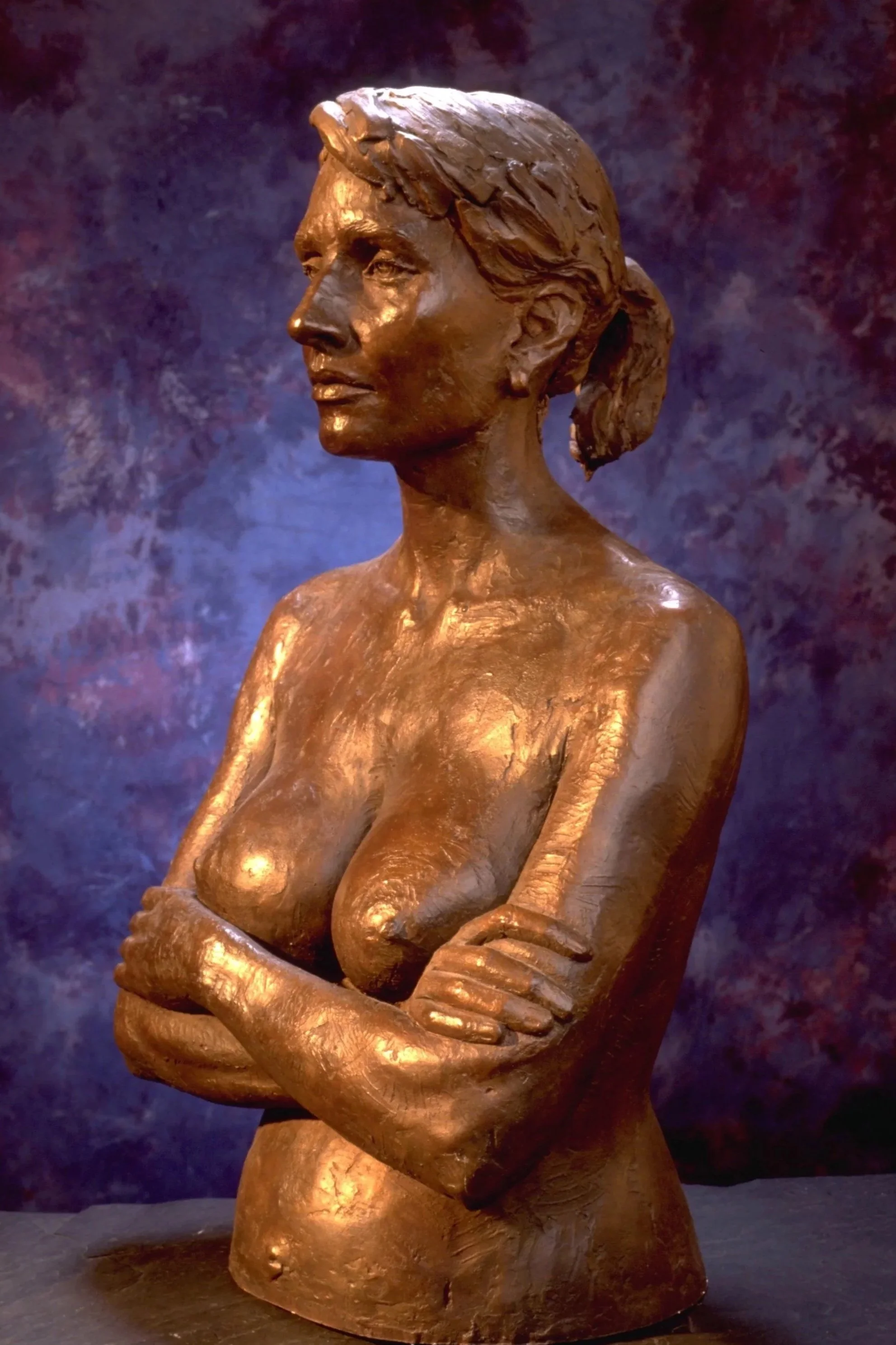 Bronze sculpture of a woman with crossed arms, with her hair tied back, against a dark patterned background.