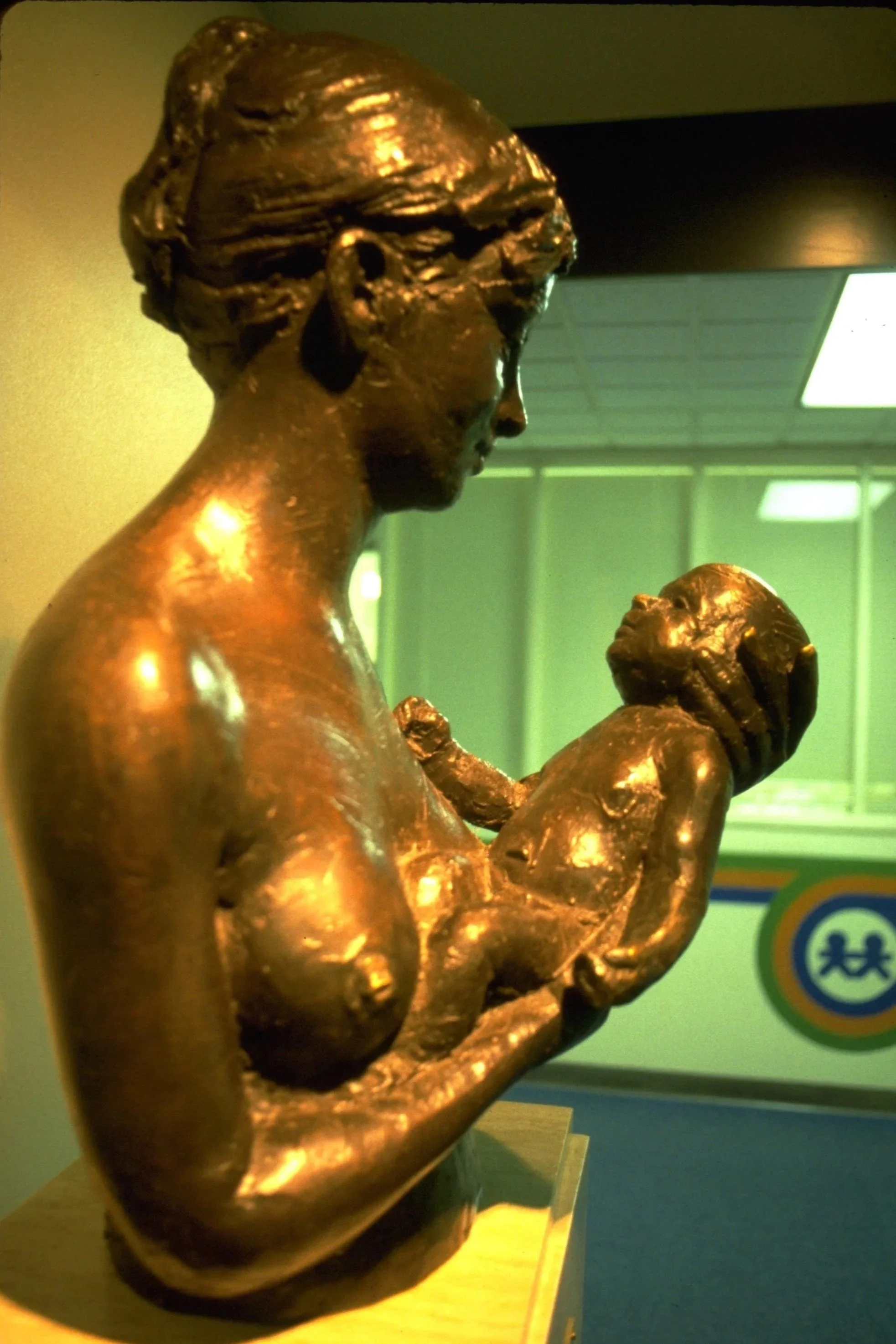 A bronze sculpture of a woman holding a small child close to her chest. The woman has short hair and is looking downward, while the child appears to be looking up. The sculpture is displayed indoors, on a wooden stand, with a blurred background showi