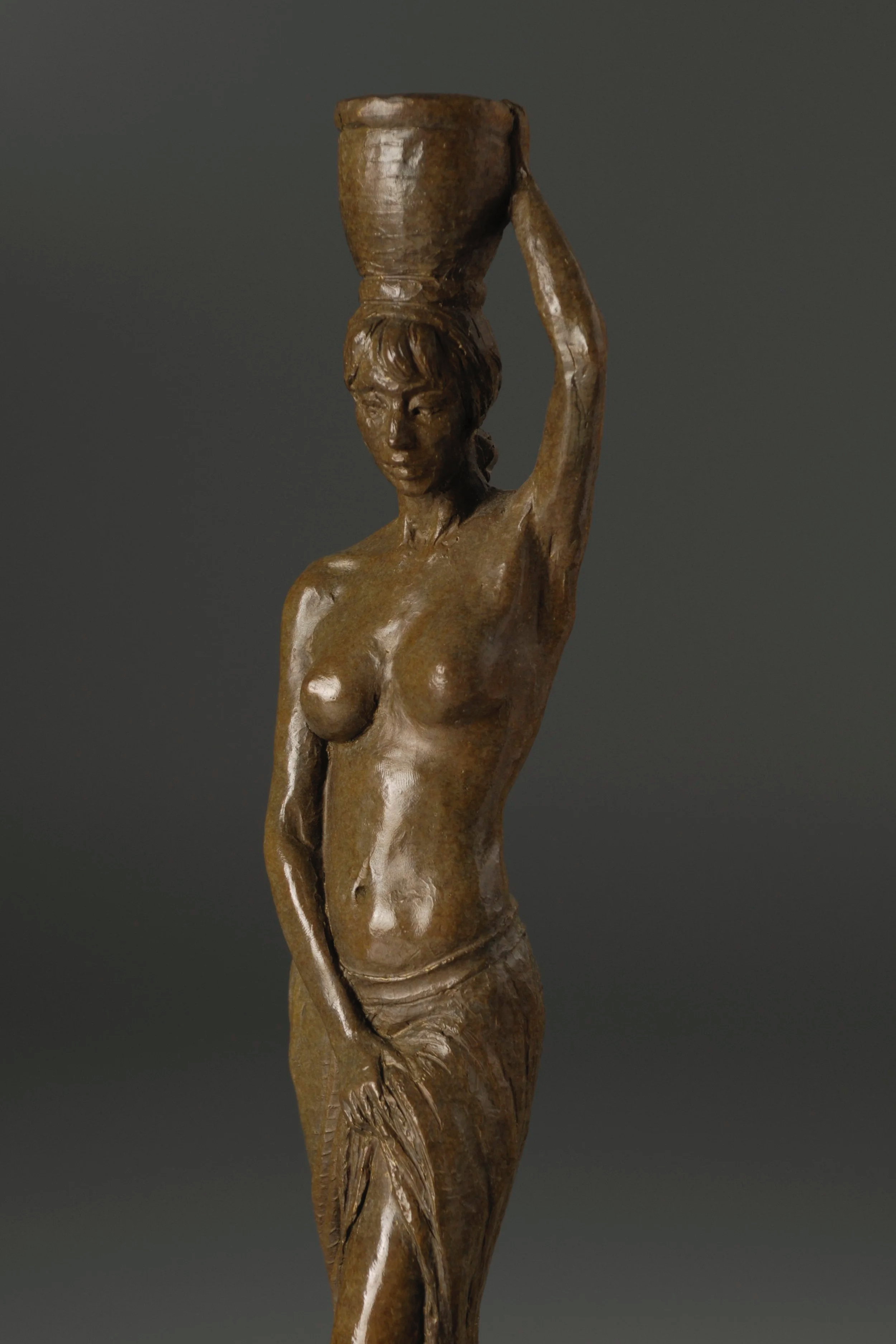 A bronze sculpture of a woman balancing a large urn on her head, with detailed features and a sleek finish, against a dark background.
