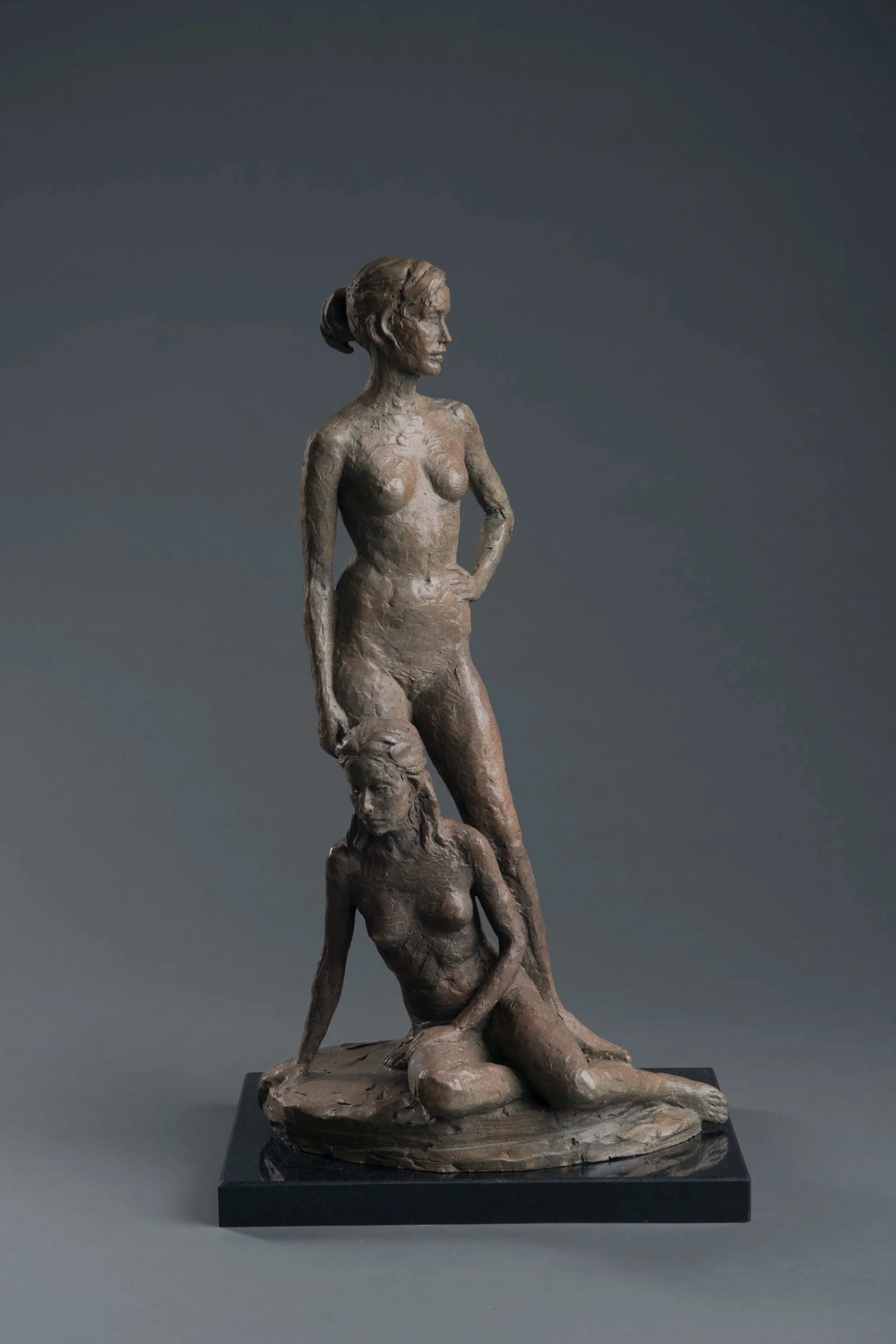 Bronze sculpture of a woman standing on a rock with another woman at her feet, on a black base, against a dark background.