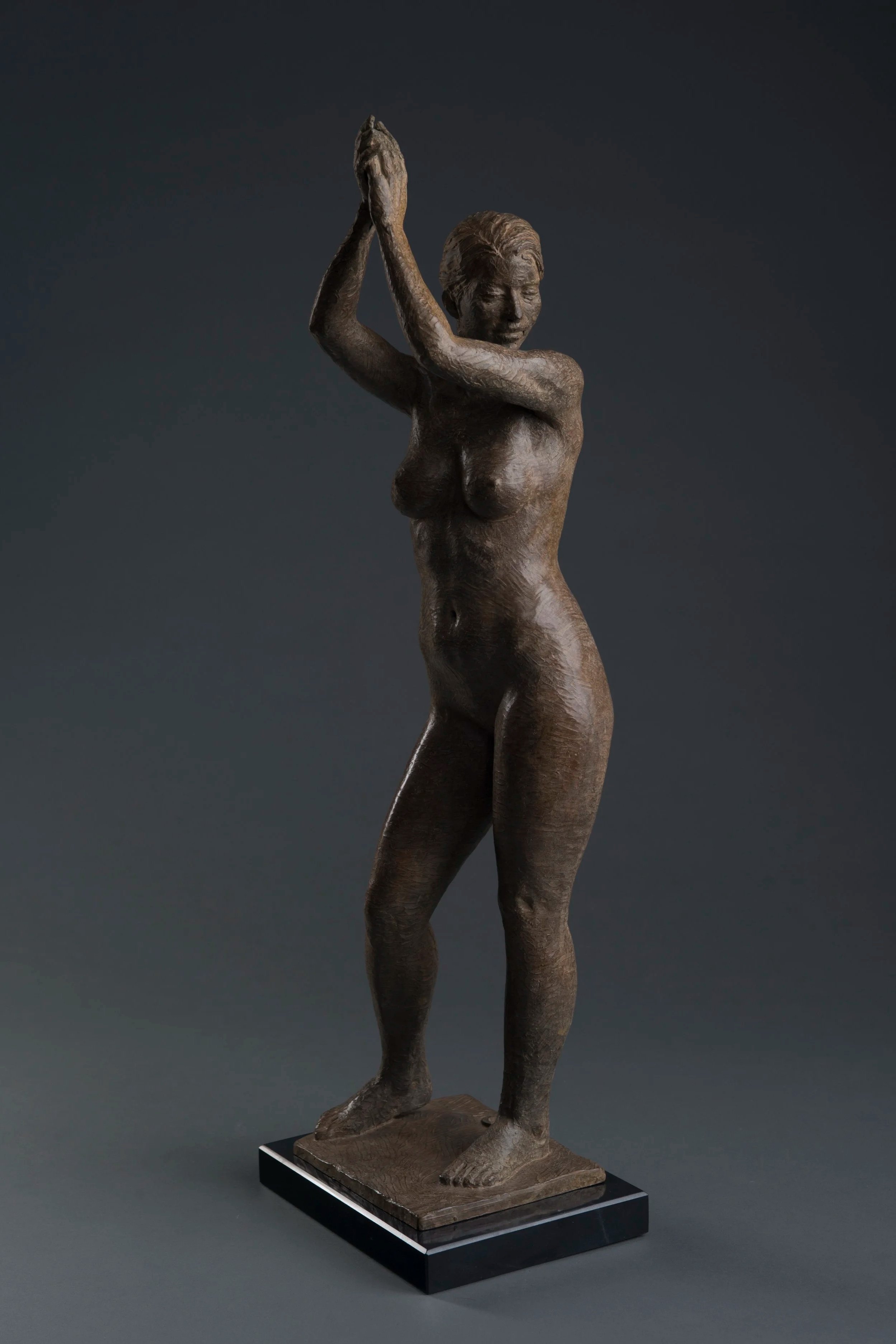 Bronze sculpture of a woman with short hair, standing with one leg slightly bent, arms raised above her head with palms pressed together, on a black rectangular base.