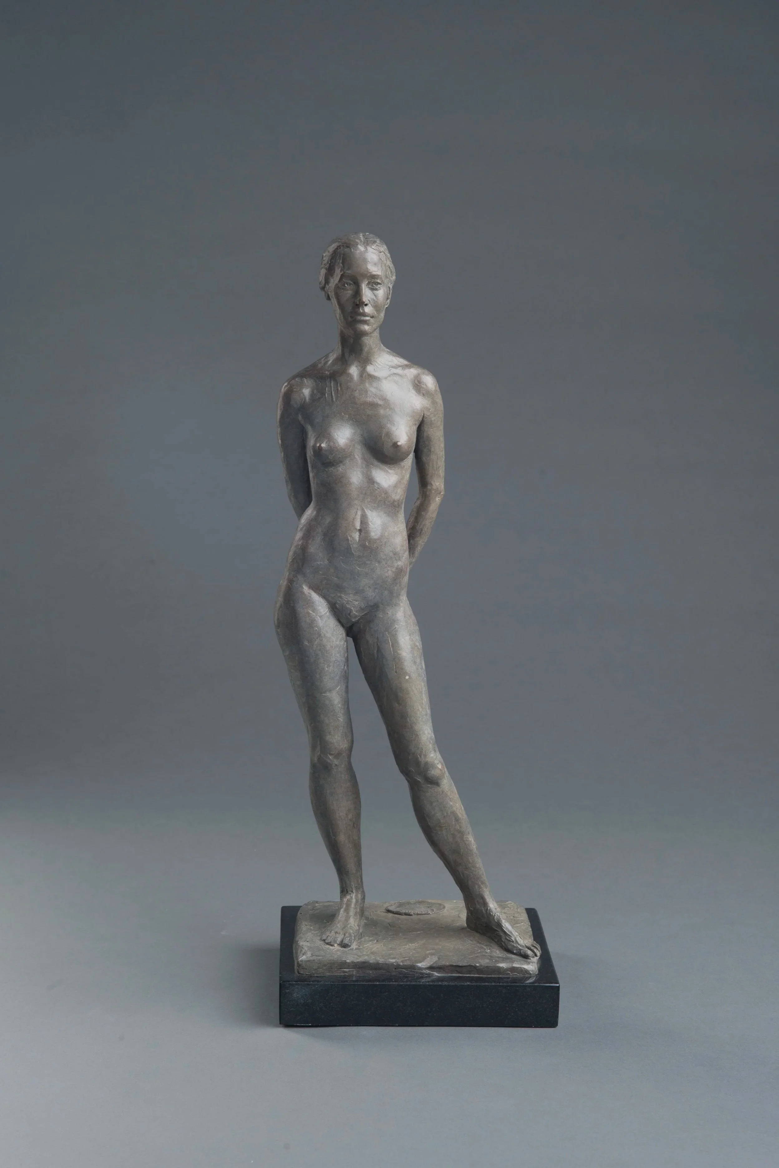 Bronze sculpture of a standing woman with short hair, one leg slightly bent, her arms behind her back, on a black rectangular base.