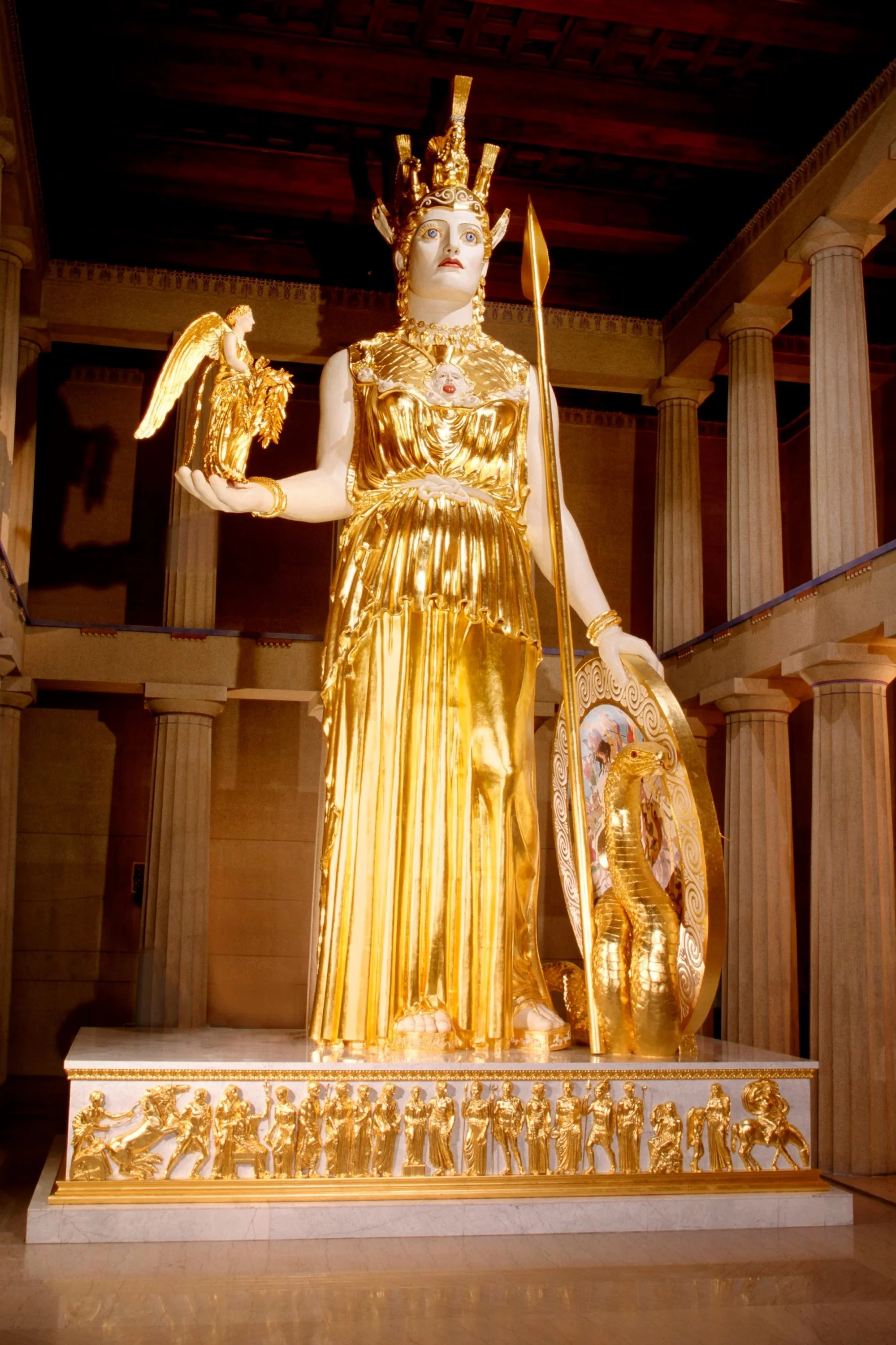 Athena Parthenos in the Nashville Parthenon by Alan Lequire.  adorned with a crown and jewelry, standing on a decorated platform with a row of smaller gold figures at the bottom, in a classical-style hall with columns.