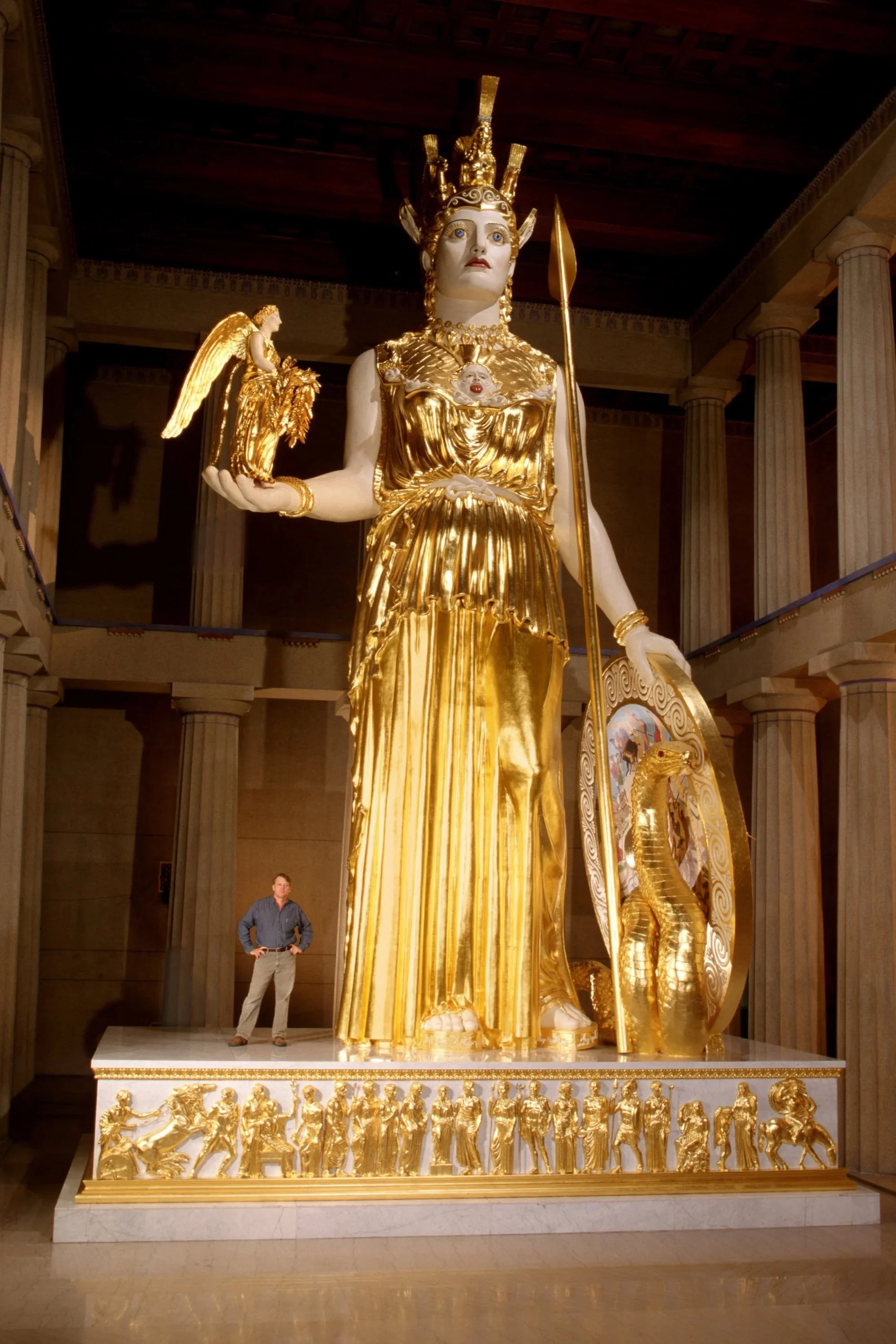 A large golden statue of a Greek goddess holding a smaller statue of an angel with golden wings. The goddess is dressed in flowing gold robes and has a golden crown. The base of the statue features detailed golden reliefs of human figures. Athena 